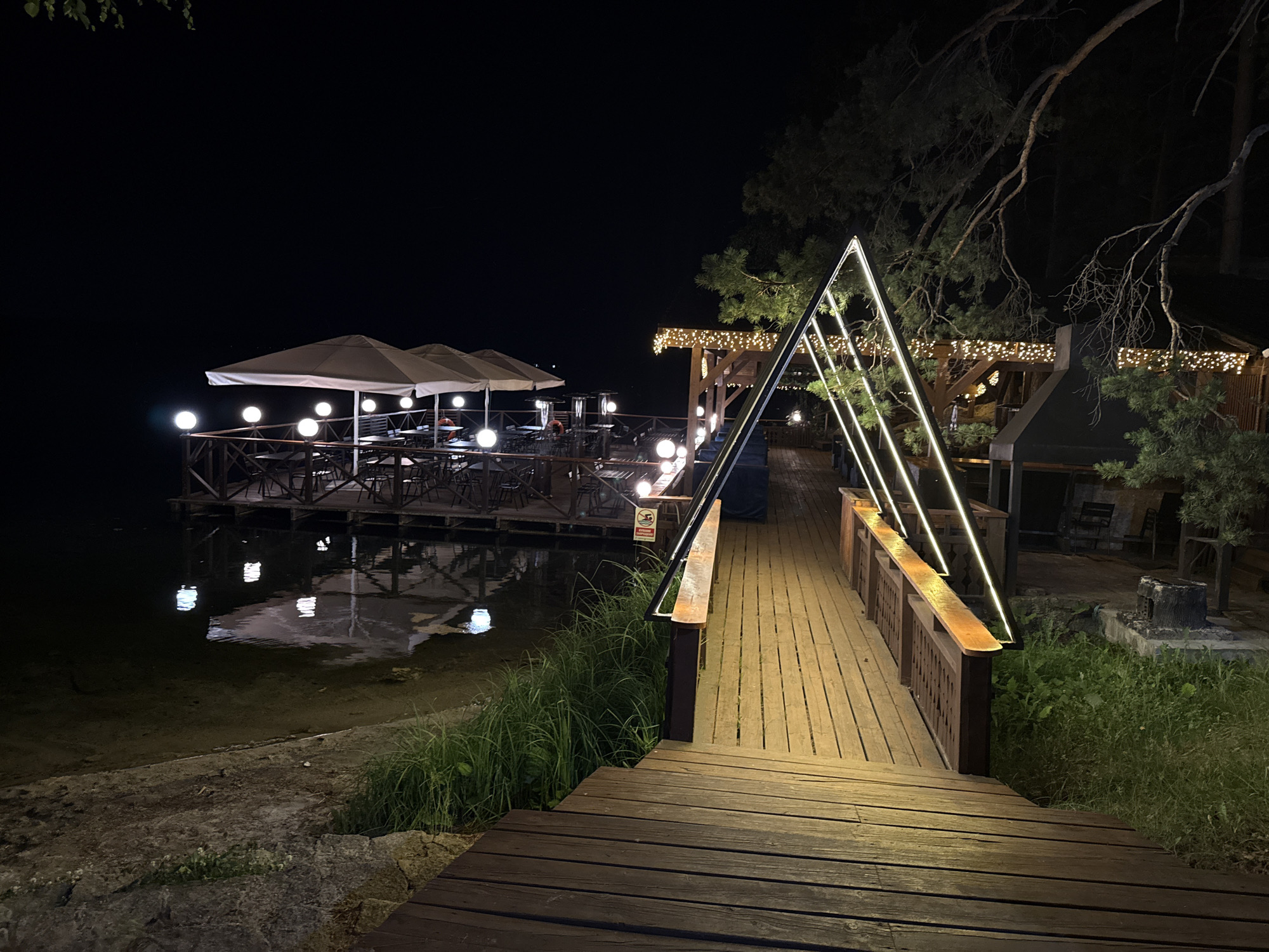 An outdoor dining area is illuminated at night by various lights. A wooden pier leads to a floating restaurant or deck situated on a body of water. The deck is covered with umbrellas and tables and chairs. String lights and triangular light fixtures adorn the area, casting a warm glow. Lush green grass and trees surround the scene, with the dark sky serving as a backdrop. The location is identified as Zolotoi Plyazh, Russia.