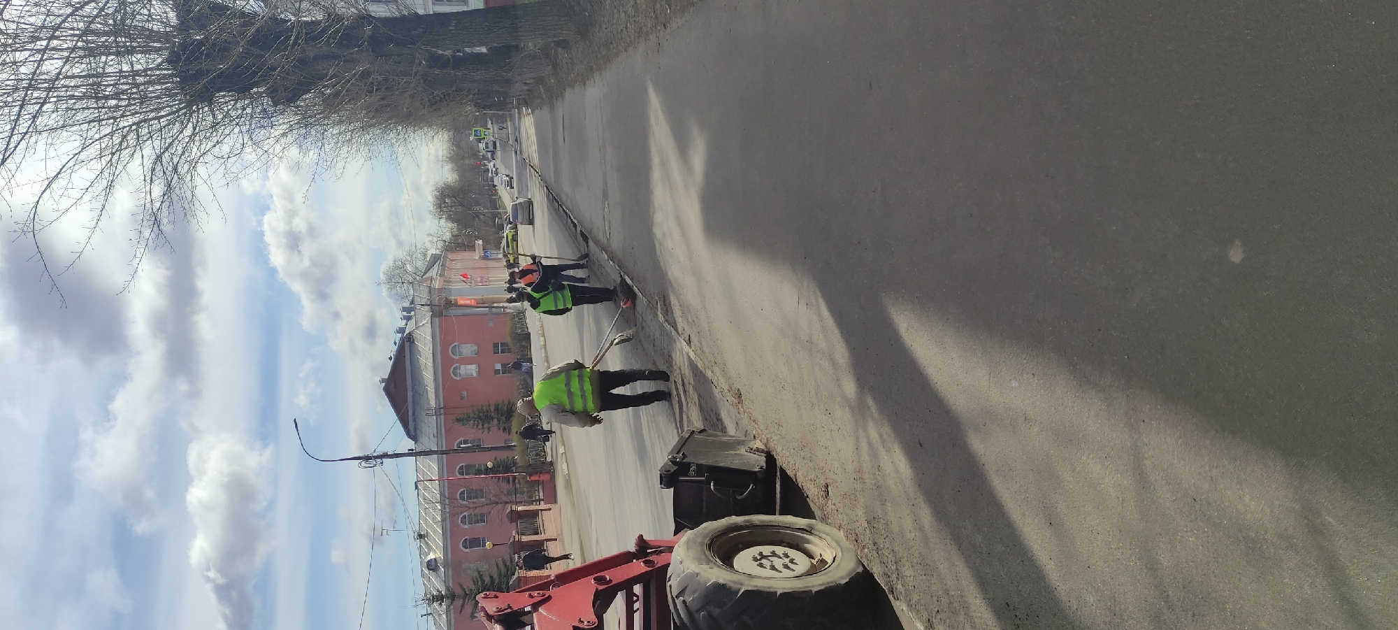 In Fryazino, Russia, a street cleaning crew is working on the side of a road. The crew members wear bright yellow safety vests. One worker is using a broom, another seems to be using a shovel. A red tractor is parked nearby. A red and pink building with a few visible stories is in the background. Above, the sky is partly cloudy. Bare tree branches are visible along the top.