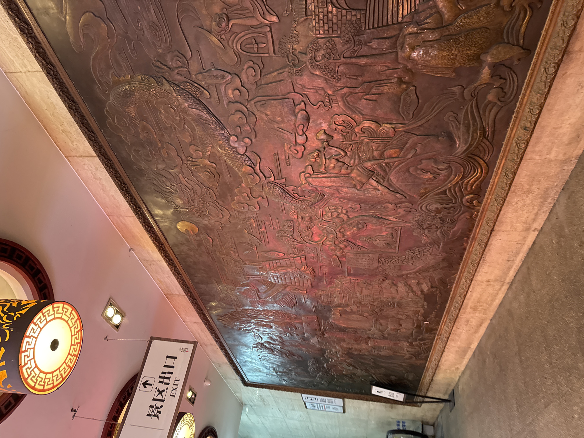 An immense, ornately framed bas-relief mural, likely crafted from bronze or a similar metallic material, dominates the wall of a seemingly public indoor space, possibly a corridor or lobby. The mural depicts a complex, layered scene rich with traditional Chinese imagery. At the top, a large, stylized dragon coils amidst swirling clouds, its form detailed with scales and a fierce expression. Below the dragon, the mural transitions into a narrative landscape populated by numerous figures, pagodas, buildings, trees, and mountains. Several figures are engaged in activities: one appears to be playing a wind instrument, others are depicted in procession or interacting within architectural structures. The colors of the mural are predominantly warm, ranging from deep browns to burnished coppers, with highlights catching the light to accentuate the three-dimensional elements. The lighting in the space is a mix of warm incandescent and cooler fluorescent tones. Above the mural, a modern white sign with Chinese characters and the word "EXIT" is visible, indicating an egress point. The floor in the foreground is a mottled gray concrete, and a portion of an escalator can be seen on the far left. The overall impression is one of cultural artistry and historical narrative integrated into a contemporary environment. The location in Yuzhong District, China, suggests a setting within a city known for its historical and cultural significance.