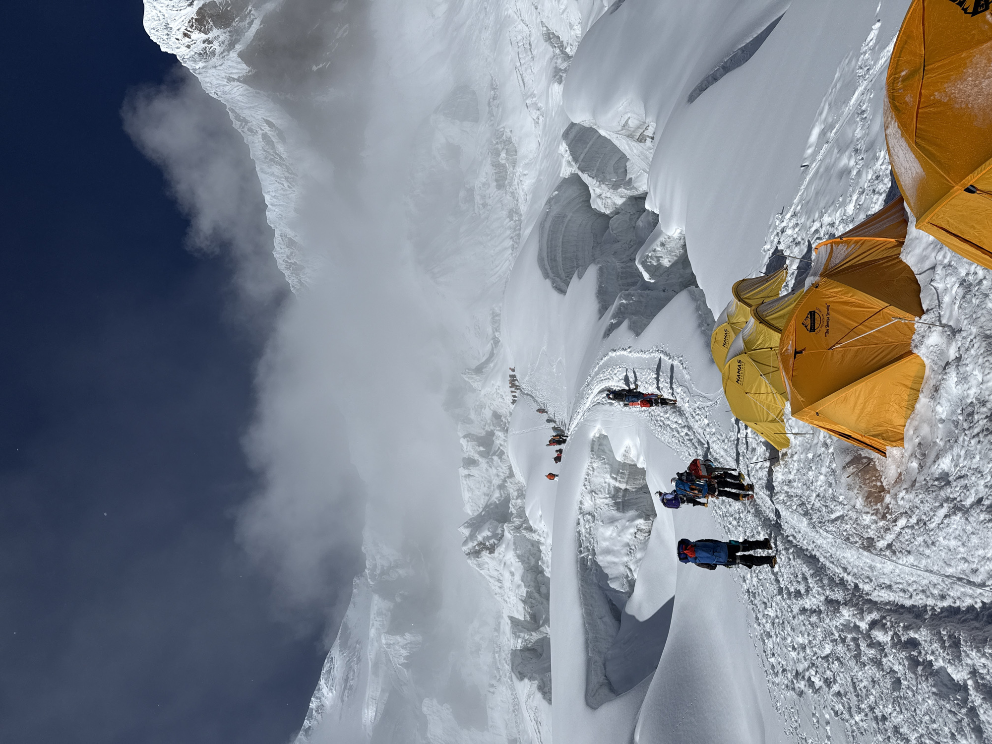 A group of mountaineers, equipped with backpacks and wearing climbing gear, ascend a snow-covered mountain trail. The trail is marked with ropes and footprints, indicating a well-traveled path. Several yellow tents are pitched along the side of the trail, some with brand names like "NAMAS" and "EXPEDITIONS" visible. The environment is a harsh, icy landscape with massive snow formations, crevasses, and towering snow-capped peaks in the background. Clouds partially obscure some of the higher mountain ranges, creating a dramatic and atmospheric scene. The sky is a deep, clear blue, with a few small white specks that could be stars or dust particles. The overall impression is of a challenging expedition in a remote, high-altitude region, likely the Himalayas, given the context of Samagaon, Nepal.