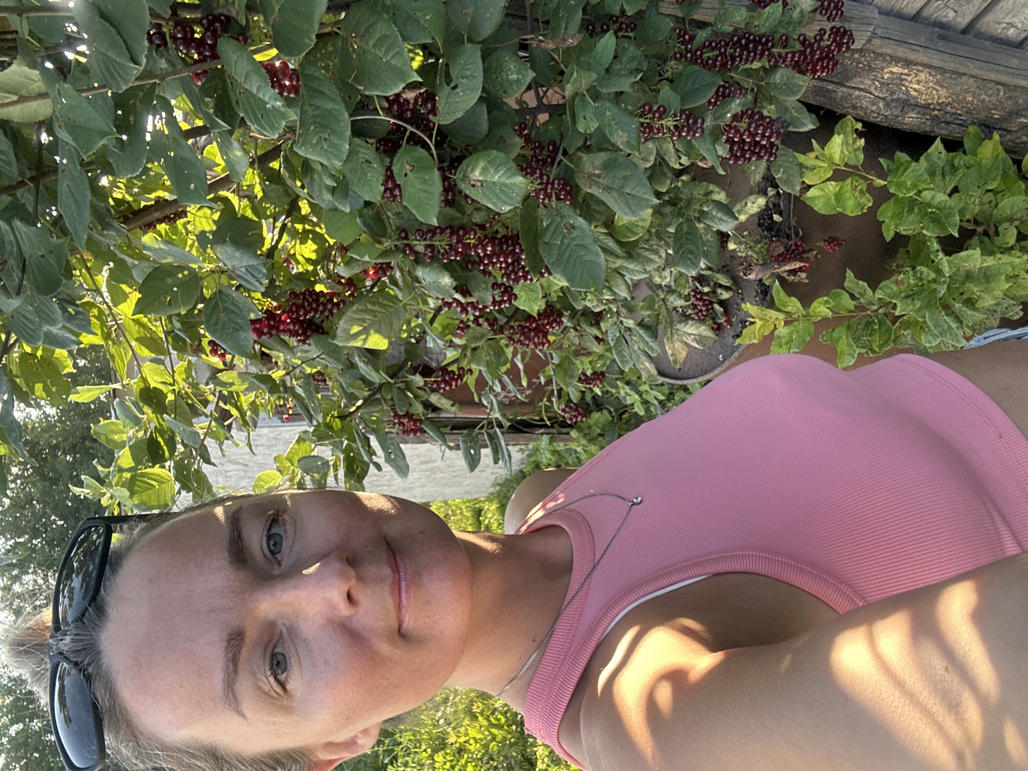 In the city of Rasskazovo, Russia, a woman with her hair pulled up in a bun, wearing sunglasses on her head, and a pink tank top, is taking a selfie. She is standing next to a plant with clusters of small, dark red berries and green leaves. The sun is shining, casting shadows and illuminating the scene. Behind her, a building with a white exterior and some weathered wooden elements are visible. The overall setting appears to be an outdoor space, possibly a garden or yard, with abundant greenery.