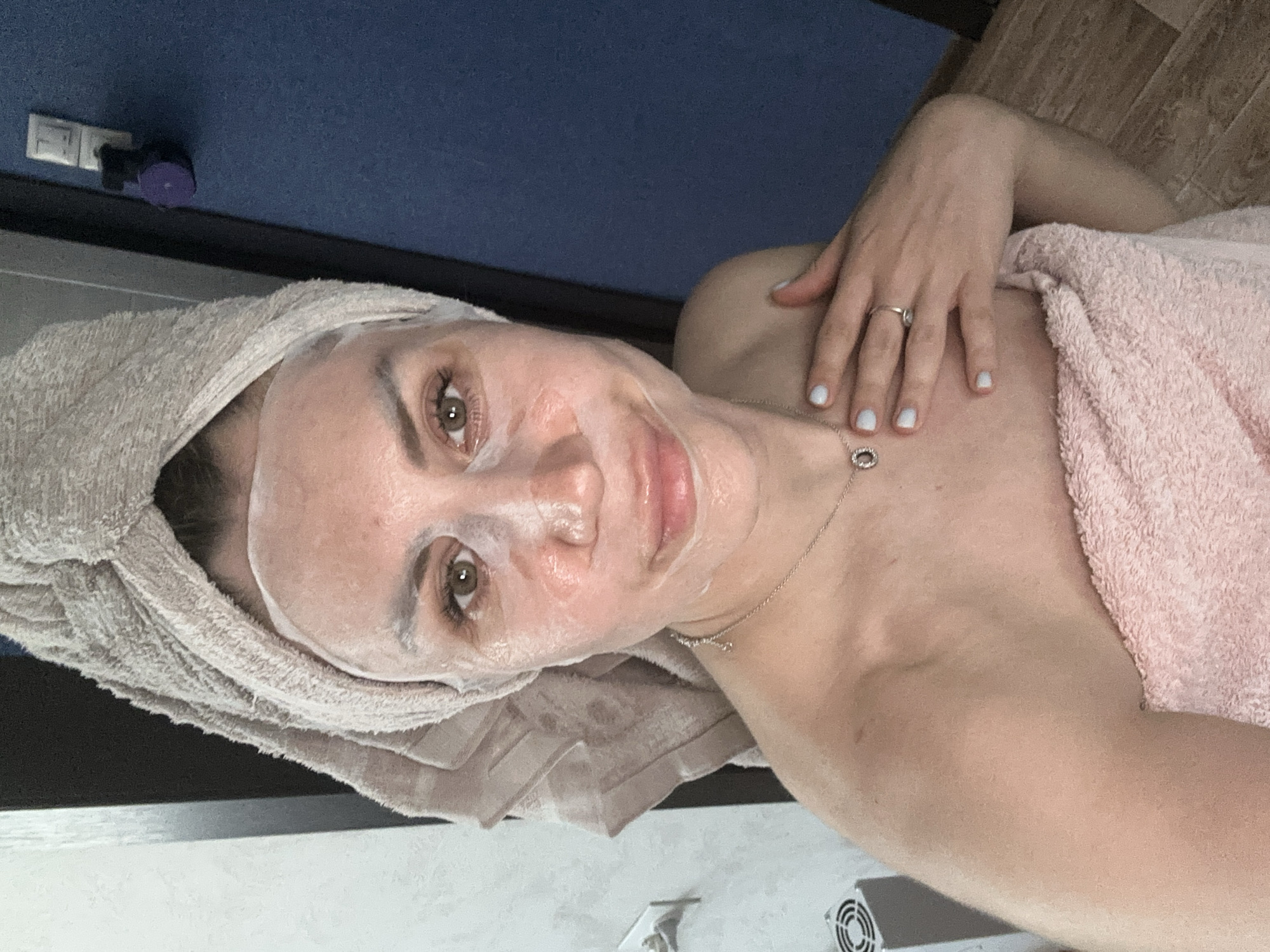 A young woman with a towel wrapped around her head and another pink towel draped over her chest is applying a sheet mask to her face. Her eyes are open and looking at the camera. She is wearing a silver necklace and a ring on her left hand. The background shows a wall with electrical outlets and a blue door. The scene appears to be in a bathroom. The image does not contain any information that would allow for its localization to Novoaltaysk, Russia.