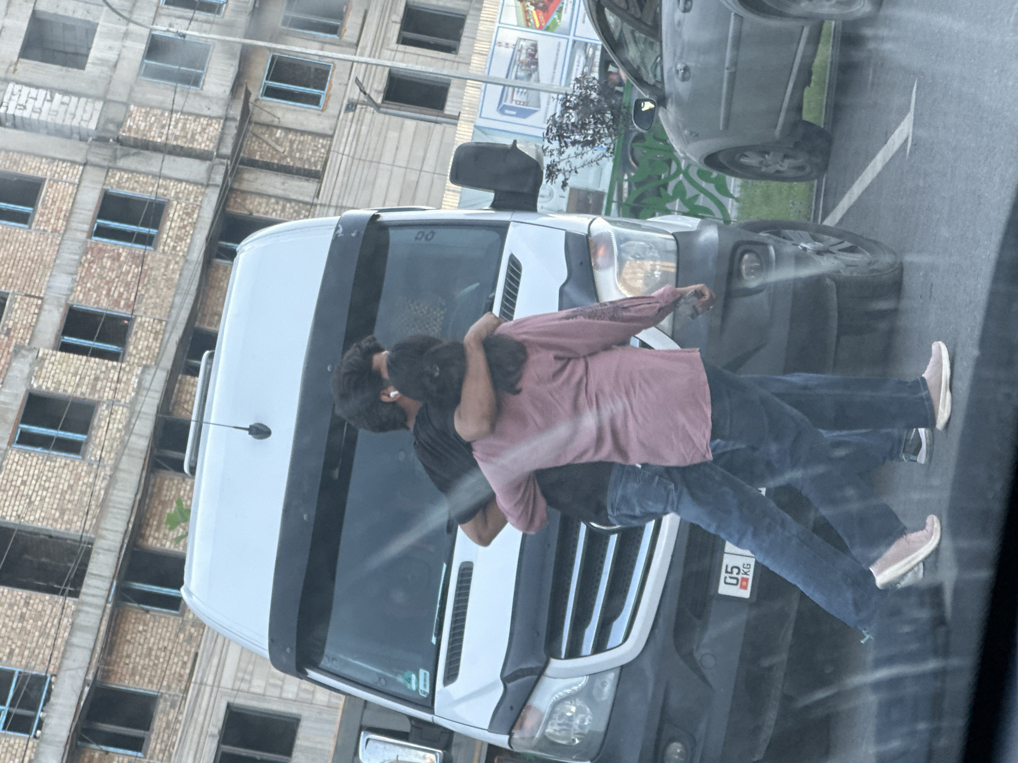 The media file shows two individuals, a man and a woman, embracing each other in the street. The man is wearing a black t-shirt, and the woman is wearing a pinkish-purple shirt and blue jeans. They appear to be standing in front of a white van, with their faces obscured. The embrace suggests a moment of affection or comfort.
The scene is set on a street with a large, multi-story building under construction in the background, characterized by exposed brick and incomplete window frames. To the right of the van, another vehicle, possibly a grey SUV, is visible. The ground is asphalt, with a white arrow painted on it, indicating a directional marking on the road.
The lighting suggests it might be daytime, perhaps late afternoon or early evening, given the relatively soft light and shadows. There are no strong indicators of specific weather conditions.
A small white license plate with red text "05 KG" is visible on the back of the van, providing a location cue. The overall atmosphere seems to be one of intimacy amidst a busy, urban environment, possibly indicating a personal moment shared between the individuals. The presence of the construction site suggests the location is an area undergoing development.