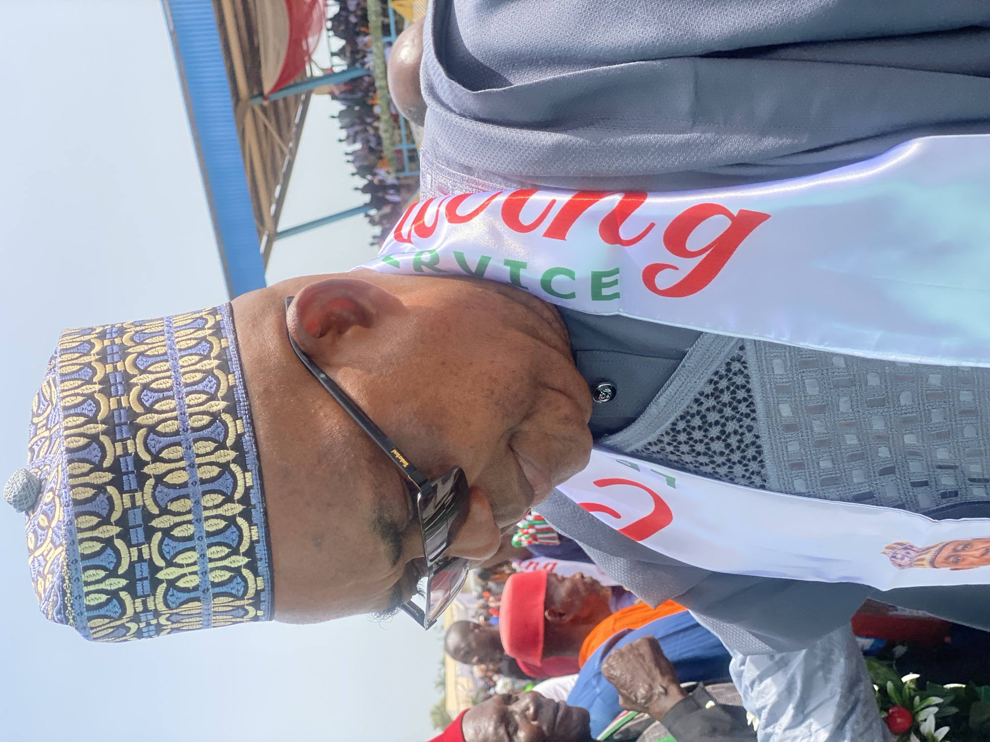 A low-angle shot captures a man wearing a traditional embroidered cap and dark glasses. He is also wearing a white sash with red and green lettering. He is positioned among a crowd of people, many of whom are looking up at him. The location appears to be an open area, possibly a stadium, judging from the overhead structure in the background. The image appears to be taken in Maigizo, Nigeria.