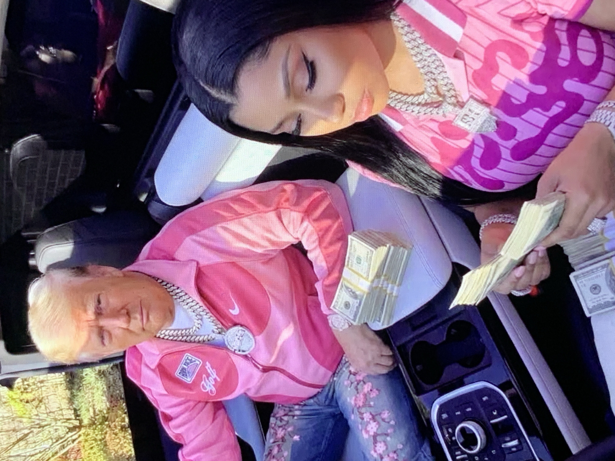 In a vehicle, two individuals are seated, surrounded by stacks of U.S. currency. On the left, Donald Trump is wearing a pink jacket and jeans adorned with a cherry blossom pattern. He has a large silver necklace on. To his right, Nicki Minaj is dressed in a pink and white striped top with a pink graphic design. She is also wearing a large silver necklace and bracelets. Both individuals are holding or near stacks of money. The vehicle's interior is visible, including a dashboard with controls and cup holders. The background suggests an outdoor setting with foliage. The overall scene appears to be a staged photograph with a focus on wealth and celebrity.