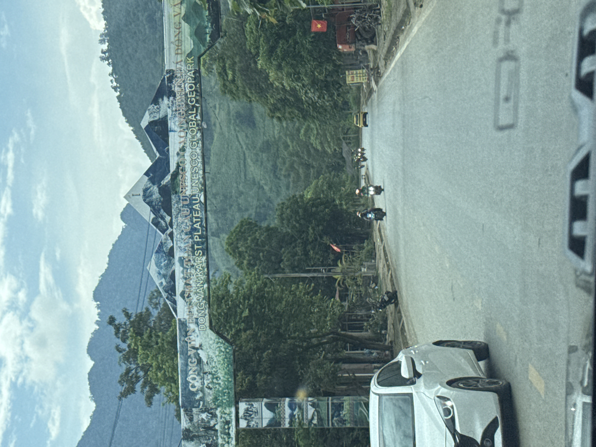 The image depicts a road in Minh Tân Commune, Vietnam, as seen from within a vehicle. A sign for the "Dong Van Karst Plateau UNESCO Global Geopark" is visible. Several motorbikes are traveling along the road, along with at least one car. Trees and mountains form the backdrop. A Vietnamese flag is also visible.