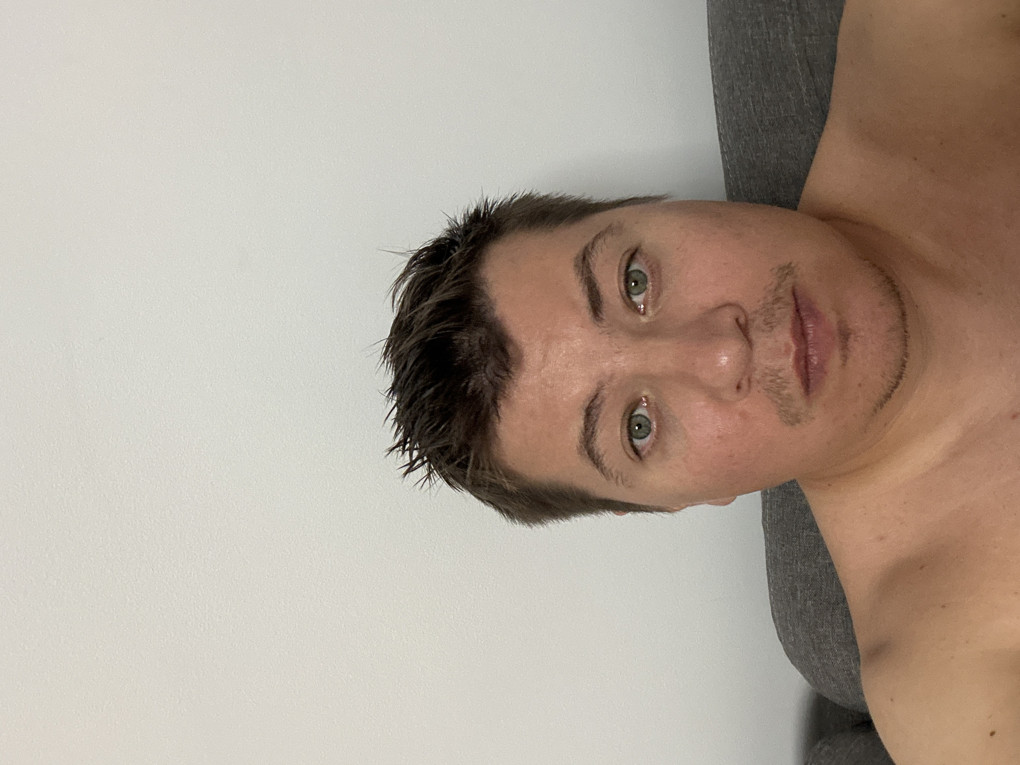 A man is photographed from the chest up, against a plain white wall. He has short, dark, messy hair and light green eyes. His skin is fair with some redness on his cheeks. He has a thin mustache and a small goatee. He is shirtless and appears to be sitting on a grey sofa. The scene is indoors, with no indication of location within the city of Benimàmet-beniferri, Spain.