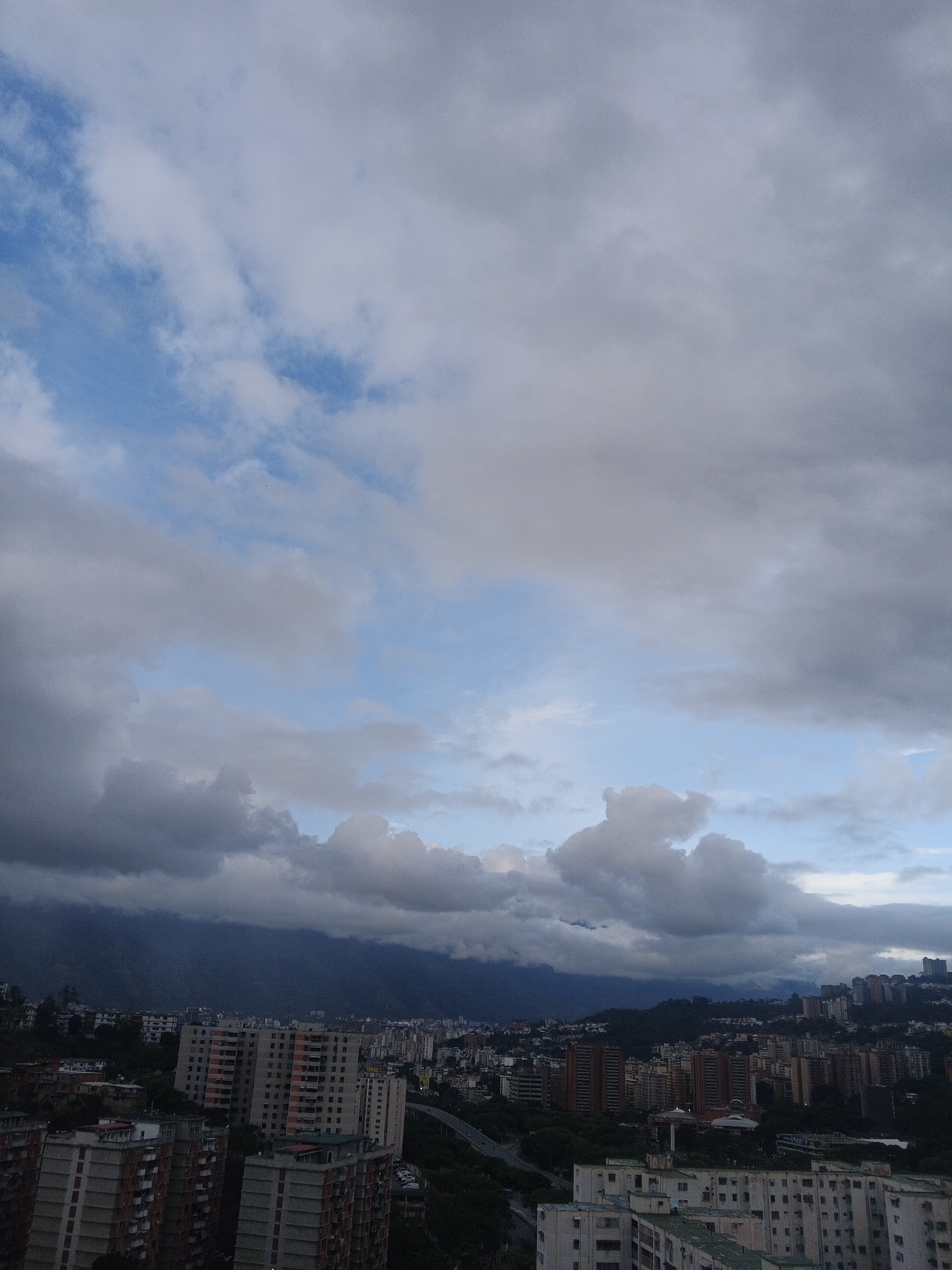 The image captures a cityscape of Caracas, Venezuela, under a cloudy sky. In the foreground and midground, dense groupings of apartment buildings dominate the scene, constructed from concrete and brick, with various heights and designs. The buildings display different shades of brown, beige, and white, showing rows of windows and balconies. A highway snakes through the city, indicating the urban infrastructure. Behind the cityscape, a mountain range can be observed, veiled by a thick layer of clouds. The sky occupies a substantial portion of the image, with alternating patches of light blue visible between layers of grey and white clouds. The overall lighting suggests either early morning or late afternoon. There is no presence of people observed in this image.