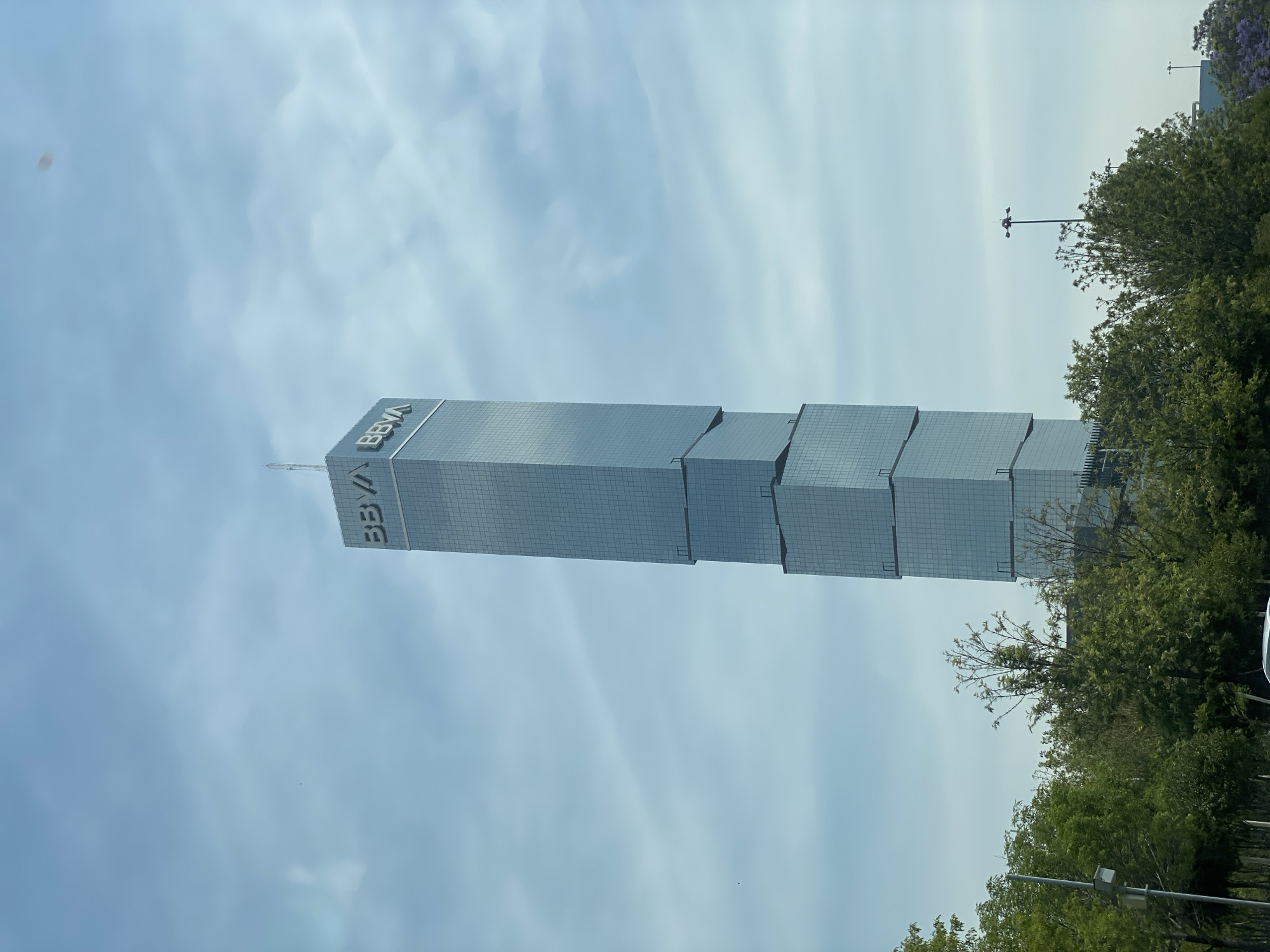 The image depicts a tall, modern skyscraper with the BBVA logo prominently displayed. The building is primarily constructed of glass and has a tiered, step-like design. To the right of the building, there is dense foliage, possibly trees, obscuring some of the structure. The sky is overcast with a light blue hue. The location appears to be an urban environment, although the specific city and country are not identifiable from the image alone.