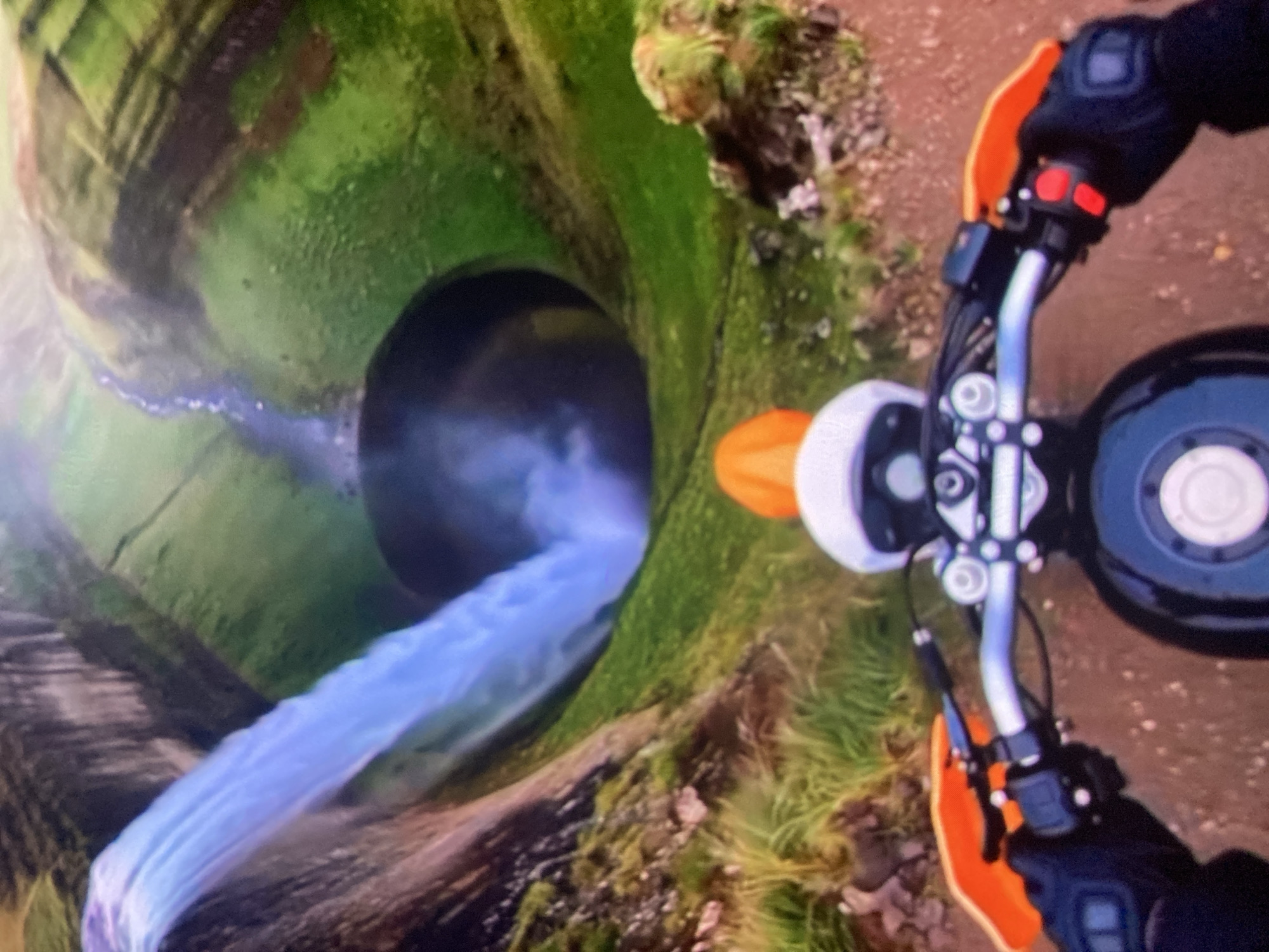 The image depicts a first-person perspective of someone riding a dirt bike. The rider is wearing black gloves with orange handguards on the handlebars. The dirt bike has a black fuel tank and a white and orange helmet is visible on top of it.
In the background, a dramatic waterfall plunges into a dark, circular abyss, surrounded by steep, green, rocky hills. A stream flows down the hillside and merges with the water of the waterfall. The environment suggests a natural, possibly remote, landscape. The image does not contain any identifiable landmarks or elements that would place it specifically in the city of Ustyvytsia, Ukraine.