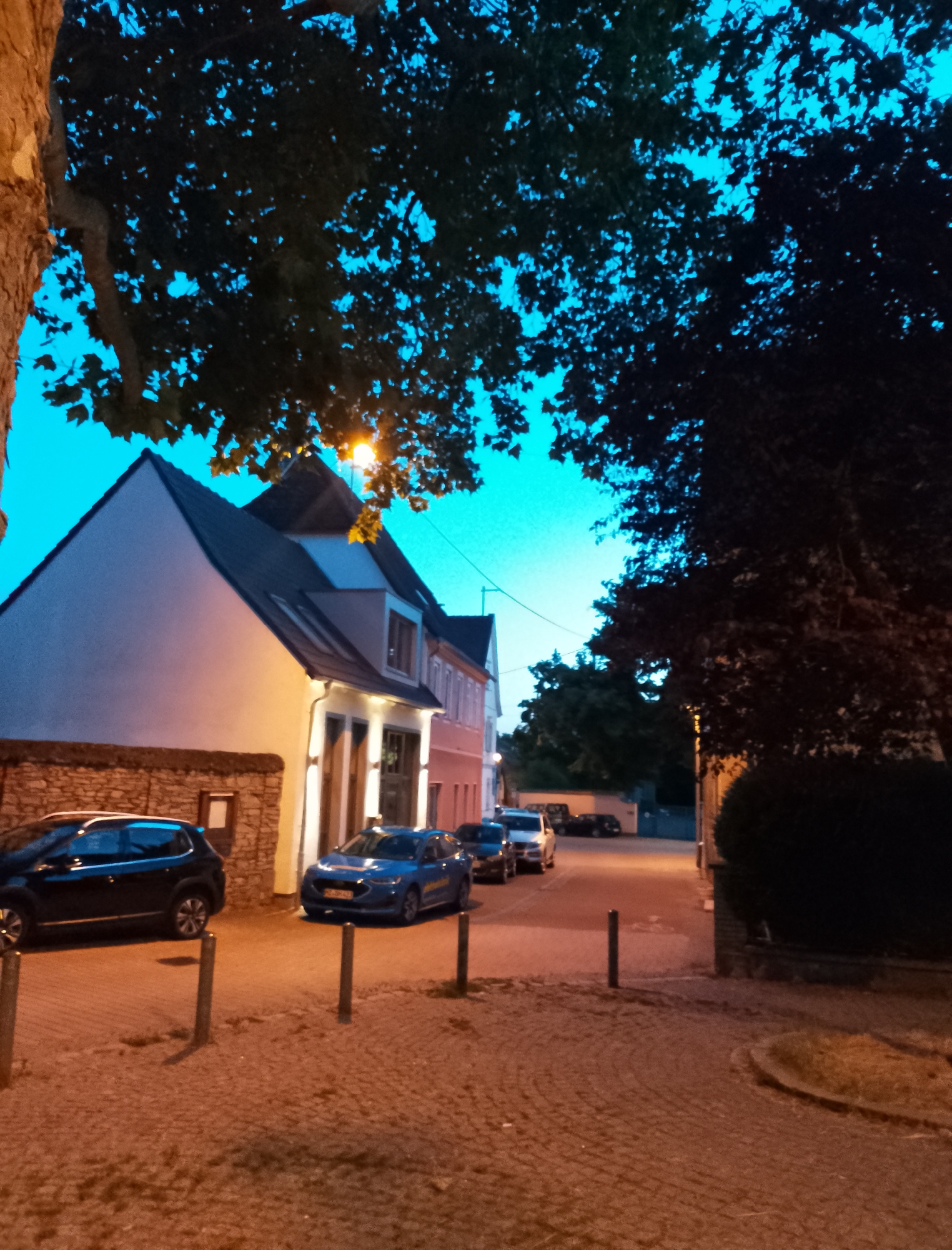 The image depicts a street scene at dusk in a European-style town. The sky is a deep, twilight blue. Large trees with dark foliage overhang the street, their leaves silhouetted against the sky. In the foreground, a cobblestone street is illuminated by the warm glow of streetlights. Several cars are parked along the street, including a dark SUV on the left and a blue sedan in the center. The buildings lining the street are multi-story, with pitched roofs and light-colored facades, some with visible windows and doors. The overall impression is one of a quiet, residential street at the end of the day.