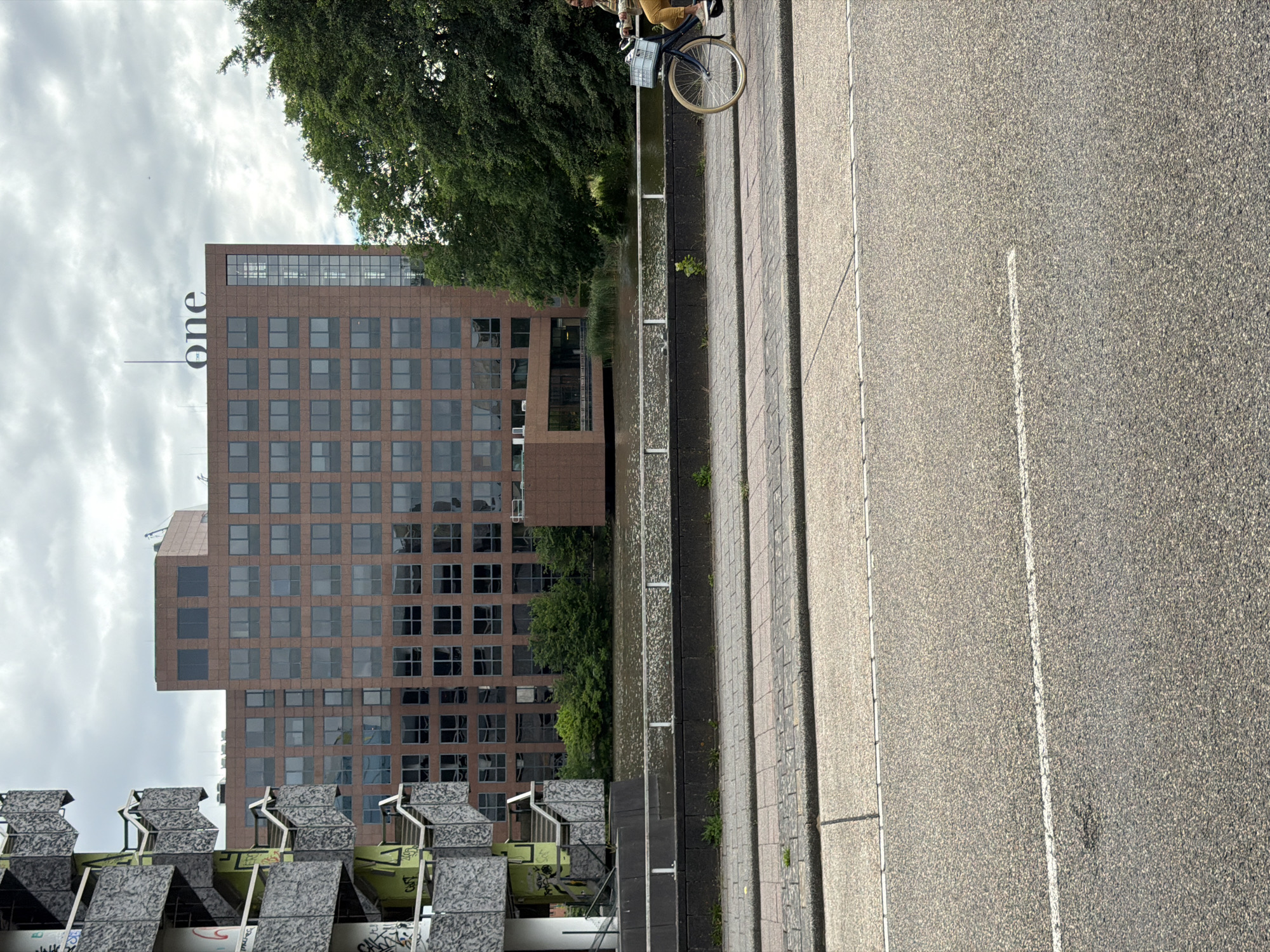 A person rides a bicycle with a light blue basket on the right side of the image, on a paved walkway. The walkway is adjacent to a body of water. In the background, a large, multi-story brown building with many windows stands. The building has the word "one" in large white letters on its upper facade. To the left of the brown building is a gray, multi-level structure with external staircases, appearing somewhat dilapidated and covered in graffiti. Trees line the edge of the water between the walkway and the buildings. The sky is overcast with clouds. The foreground shows a section of asphalt road with a white line. This scene is located along the Amstel River in Amstelveen, Netherlands, near the office building commonly known as "The One" or "De Kantoorkolos" for its distinctive appearance and the prominent signage.