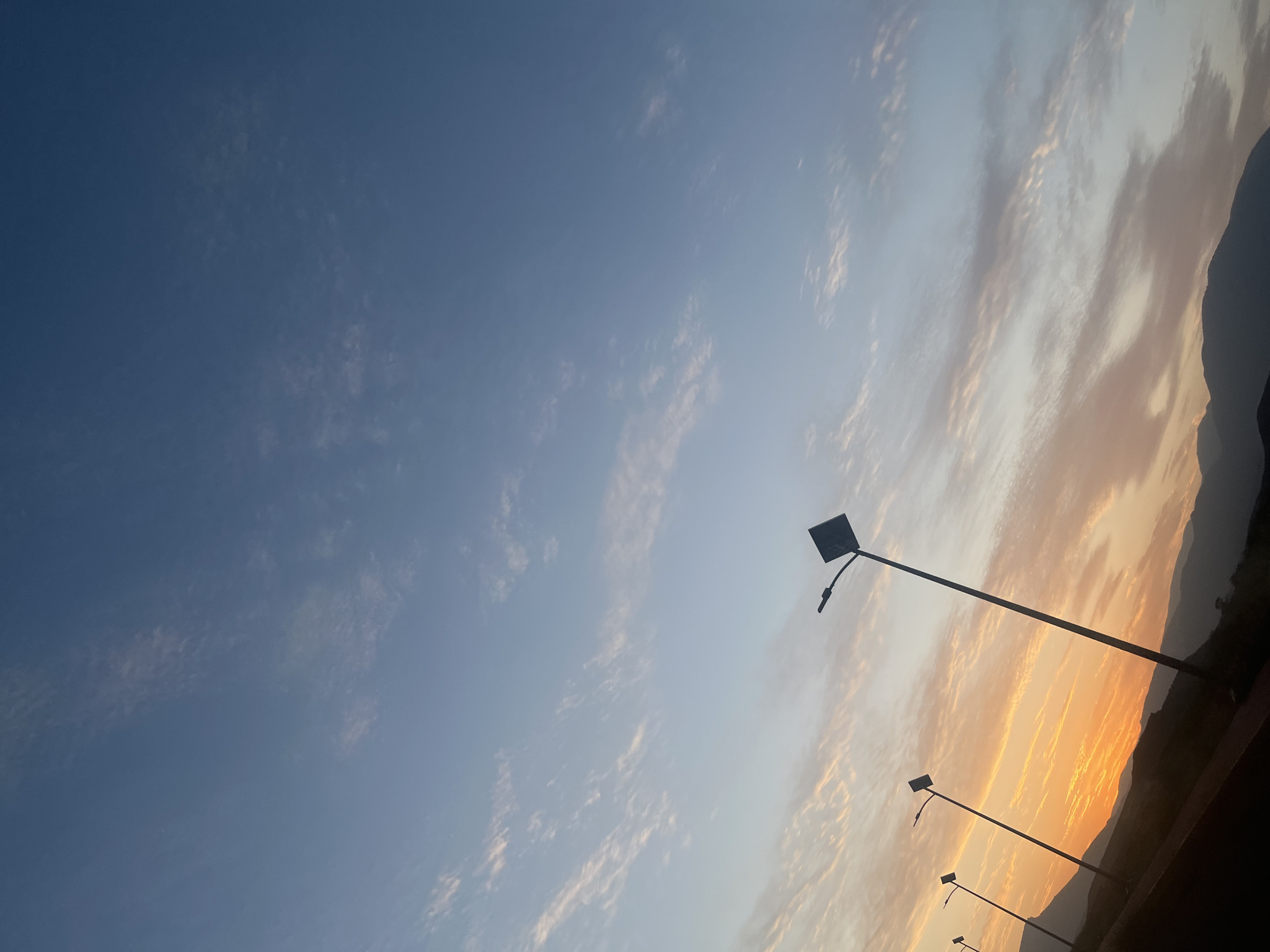 The image shows a section of Bin El Ouidane, Morocco at sunset. The sky is a gradient of blue to orange. There are clouds stretched across the sky, tinted with pinks and oranges near the horizon. The silhouette of a mountain range is visible in the far right of the frame. A row of streetlights stretches diagonally across the image, from the top right to the bottom right.