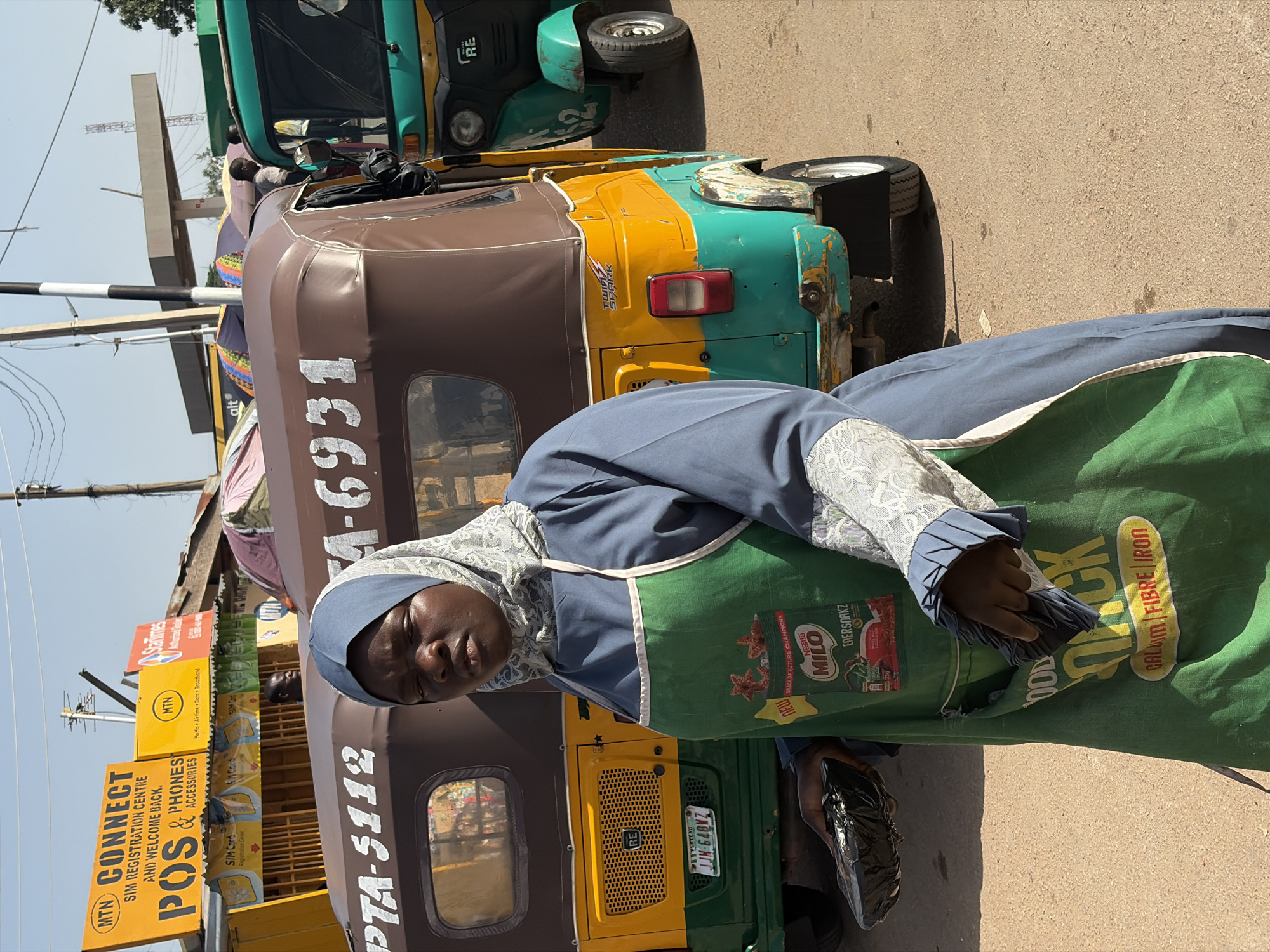 In Gura Top, Nigeria, a person wearing a head covering, a blue garment with lace trim, and a green apron with the "Milo" brand visible stands on a street. The apron also displays "QUICK FOOD" in large letters with the text "CALUM FIBRE IRON" underneath. The person is holding something dark and crumpled in their hand. Behind them, a yellow, green, and brown auto-rickshaw displays the license plate "JJN 648NZ" and the number "PTA-5112". Other auto-rickshaws are parked nearby. A sign advertises "MTN CONNECT" for "SIM REGISTRATION CENTRE", "POS", and "PHONES & ACCESSORIES". Another sign promotes "MoMo Airtime Data Broadband". A "StarTimes" sign is partially visible in the background. A utility pole stands near the signs, and the scene is captured in bright daylight. The number "4-6931" is written on an auto-rickshaw.