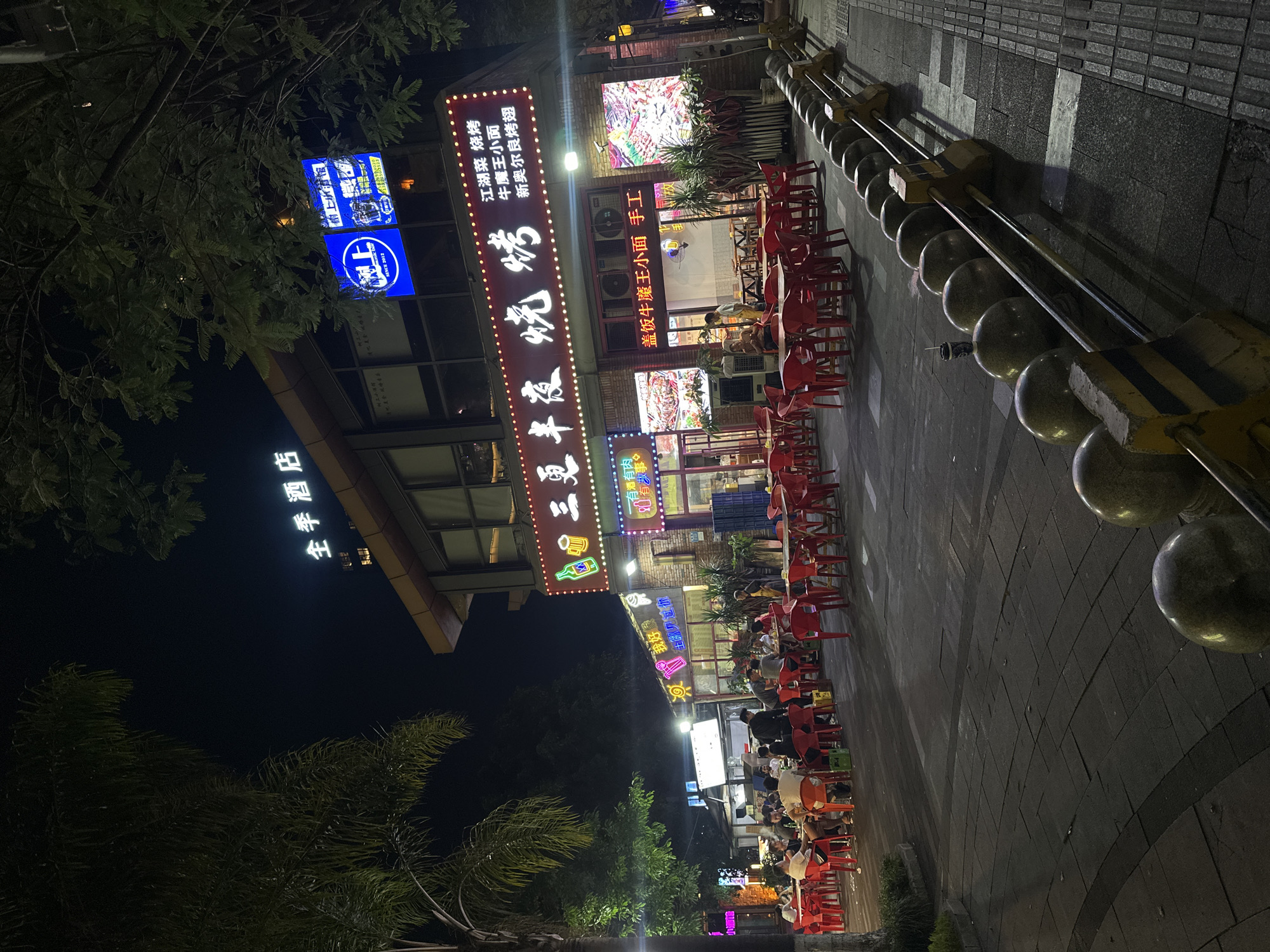 At night, an outdoor dining area is set up in front of a restaurant with a neon sign that reads "三更半夜烧烤" (Midnight Barbecue). Red plastic tables and chairs are arranged on the pavement, with several groups of people seated and dining. The restaurant facade is brick, and above it, another sign displays "全季酒店" (Hanting Hotel). Adjacent to the barbecue restaurant, another establishment features a bright blue neon sign with the logo "上" and Chinese text, indicating a business called "上伴·主题馆" (Shang Ban Theme Hall). The scene is illuminated by various neon signs and streetlights. To the right, a sidewalk with a decorative barrier of large, spherical stone bollards and metal railings separates the pedestrian area from the street. Lush green trees frame the scene, contributing to the ambiance of a bustling urban night market.