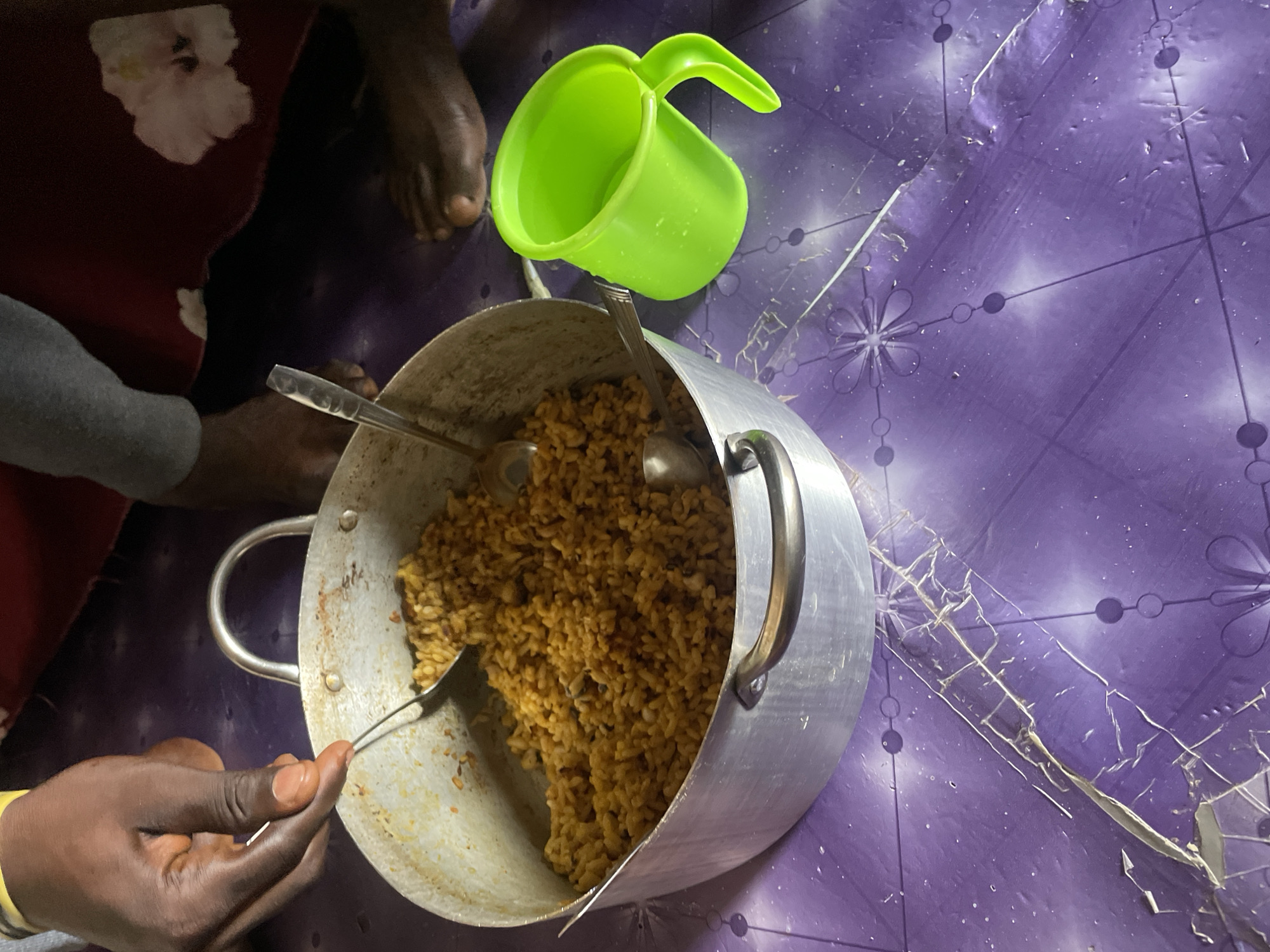 This image depicts an overhead view of a person's hands scooping rice from a large aluminum pot placed on a purple, patterned plastic mat. To the right of the pot sits a bright green plastic mug filled with water. The scene appears to be indoors, possibly in a residential setting within Abuja, Nigeria. The lighting suggests it might be daytime, but the overall exposure is somewhat dark. There are other individuals' feet visible in the background, suggesting a communal meal or gathering. The mat itself shows signs of wear and tear, with some areas appearing creased or damaged. The rice in the pot is a golden-brown color, and it is mixed with what appear to be beans or other small dark ingredients. Two metal spoons are partially submerged in the rice, one actively being used by the person.