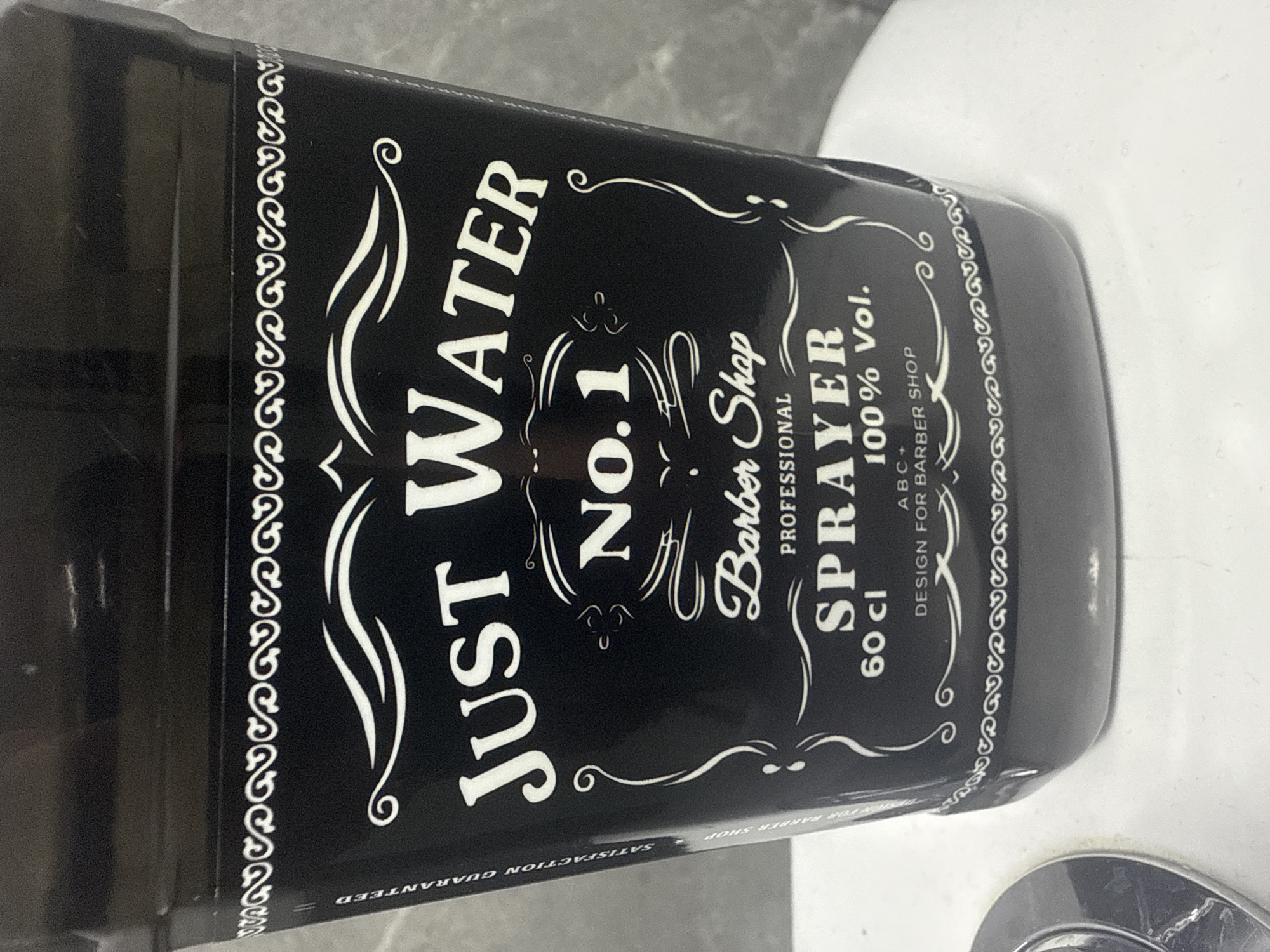 A black spray bottle with white text and decorative elements sits on a white surface, possibly a sink or countertop, with a grey, textured wall in the background. The bottle has a label that reads "JUST WATER NO. 1 Barber Shop PROFESSIONAL SPRAYER 60 cl 100% Vol. DESIGN FOR BARBER SHOP." A white decorative border runs along the top and bottom of the label. There is also text on the side of the bottle that reads "SATISFACTION GUARANTEED FOR BARBER SHOP." A portion of a silver object, possibly a faucet or drain cover, is visible in the lower left corner of the image.