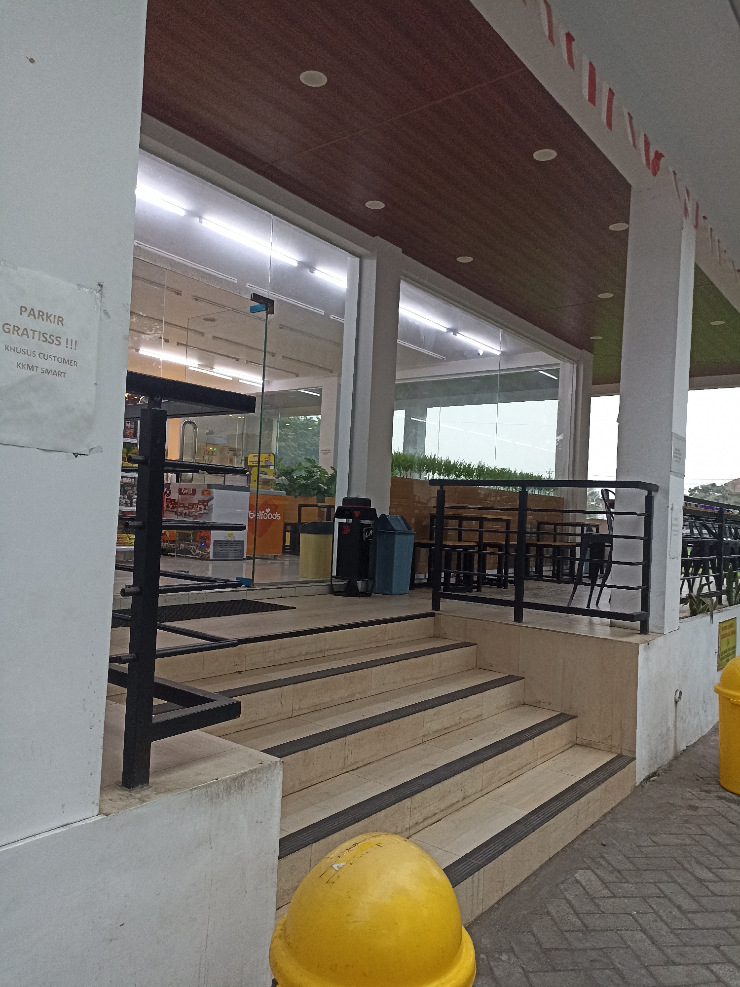 The image depicts the exterior entrance to a store, likely a convenience store or small supermarket, in Tompokersari, Indonesia. A white pillar on the left has a sign with text in Indonesian stating "PARKIR GRATISSS !!! KHUSUS CUSTOMER KKMT SMART". The entrance itself is composed of a set of beige tiled stairs with dark anti-slip strips. These stairs lead up to a glass-enclosed storefront. Inside, rows of shelves stocked with products are visible, along with refrigerators and what appears to be a cashier counter. A black metal railing lines the sides of the stairs and continues along a walkway. To the right of the stairs, there is a blue trash bin and a yellow trash bin. Beyond the immediate entrance, outdoor seating with dark metal tables and chairs is visible under a covered area. The sky is overcast.