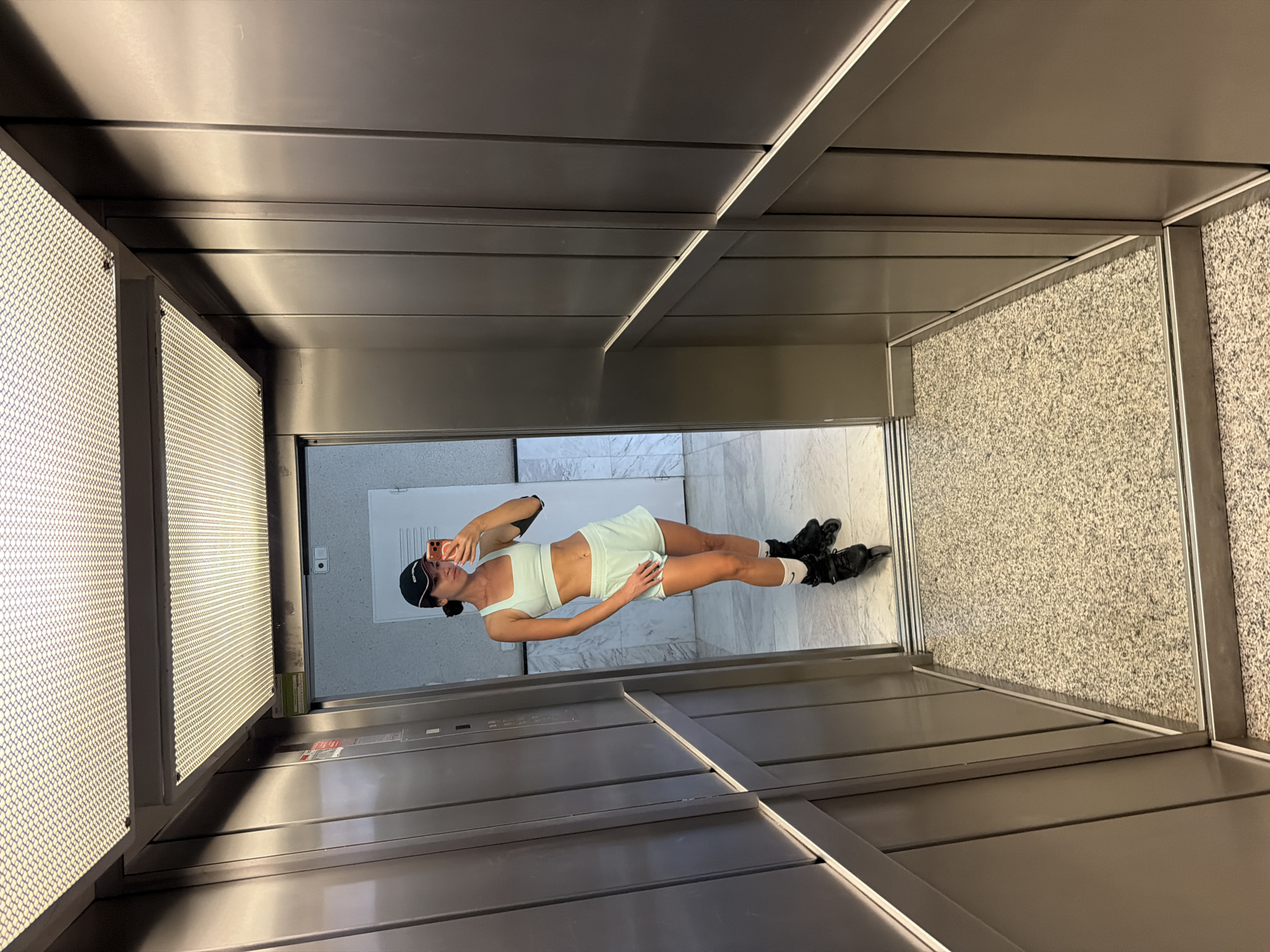 A woman wearing a mint-green sports bra and matching shorts, a black baseball cap, and black rollerblades is taking a selfie in an elevator. She is standing on a speckled gray floor, and the elevator walls are made of brushed metal. The elevator has a large mirror on the back wall, reflecting the woman and the opposite elevator wall. The ceiling has a grid of fluorescent lights. The overall impression is modern and utilitarian. The location is the city of Salou, Spain.