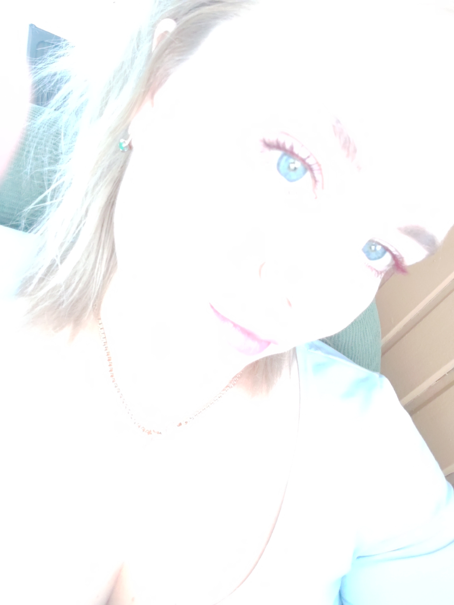 The image is a close-up, overexposed selfie of a person with blonde hair and blue eyes. The person is wearing a light blue top and a delicate necklace. Their right ear is visible and adorned with a small, green earring. The background appears to be indoors, with vertical wooden paneling visible on the right side of the frame. There is no discernible information about the location within the city of Izhevsk, Russia, nor is there any action or interaction depicted.