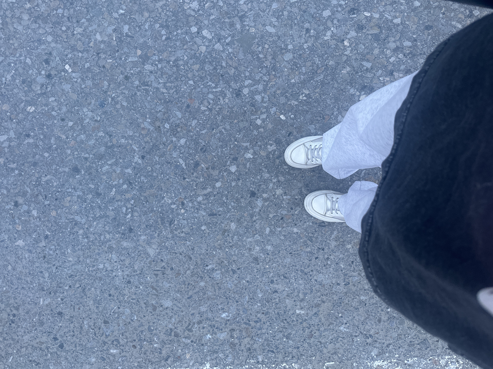 An eye-level shot shows a pair of white sneakers on a mottled grey surface, presumably asphalt, with the bottom portion of black pants on the right side of the frame. The pants partially cover a layer of light grey fabric, possibly sweatpants or leggings. The surface shows a random, speckled pattern of different shades of grey, indicative of road or sidewalk material. The location is unspecified beyond the city of Высокий, Russia.