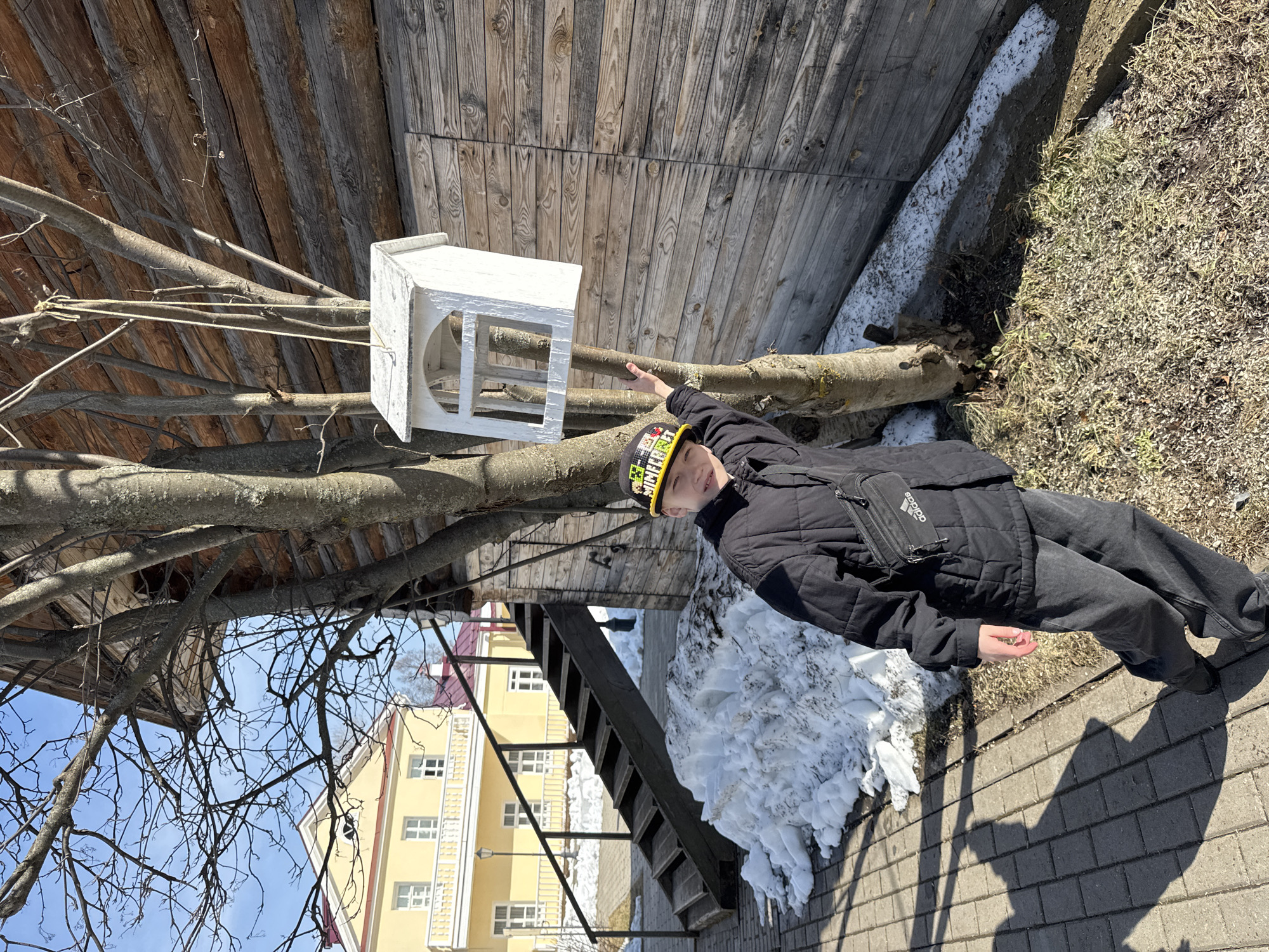 A young person stands next to a birdhouse, likely in Votkinsk, Russia. The person, wearing a black jacket, dark pants, and a colorful hat, is touching the branch supporting the birdhouse. The birdhouse is white and box-shaped. The location appears to be outdoors, adjacent to a wooden building. There is snow on the ground near some stairs. A yellow building with windows is visible in the background. The ground is paved.