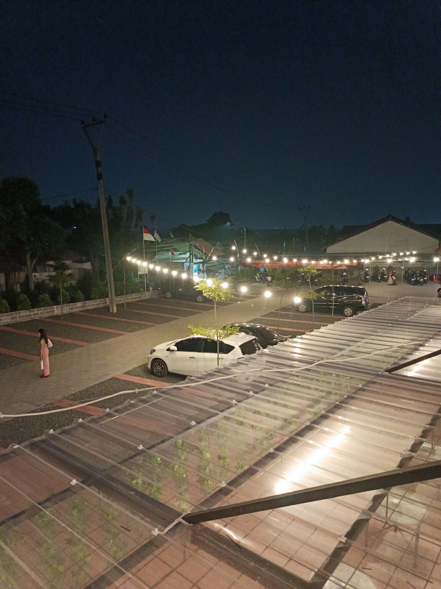 The image depicts a nighttime scene at a parking area, likely outside a commercial establishment in Cimahi, Indonesia. The sky is a deep, dark blue, indicating late evening or night. In the foreground, a transparent corrugated roof with visible structural beams is prominent, covering what appears to be an outdoor seating area or walkway with brick flooring. Below this roof, a parking lot is visible.
The parking lot contains several vehicles, including a white car parked on the left, and a black SUV and another dark vehicle further back. To the left of the parking lot, a single person, appearing to be a woman in a pink outfit, stands on a paved walkway. The walkway is bordered by gravel and brick accents.
In the background, more vehicles and people are present, suggesting a bustling atmosphere. String lights are hung across the area, illuminating the scene and casting a warm glow. Two Indonesian flags are prominently displayed, with red and white colors visible against the dark sky. Trees and utility poles with electrical wires are also part of the environment, typical of a semi-urban or suburban setting. A building with a light-colored roof is visible in the far background. The overall impression is of an outdoor dining or recreational area at night, illuminated by artificial lights and decorated with national flags.