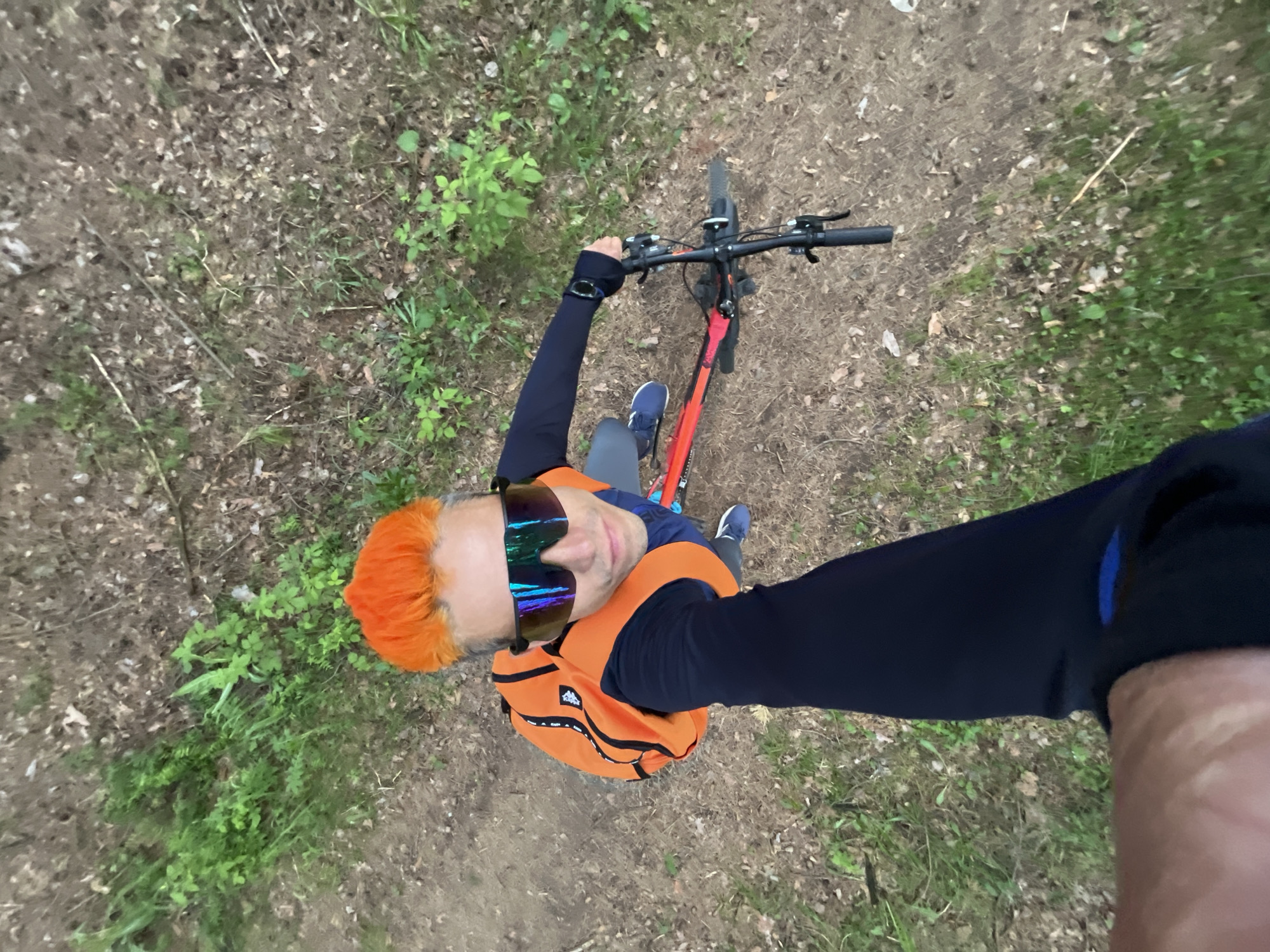 The image presents an overhead perspective of a person standing in a woodland environment. The person has bright orange hair and is wearing reflective sunglasses, a dark blue long-sleeved shirt, an orange backpack, grey pants, and blue shoes. They are positioned with a red bicycle between their legs, holding onto the handlebars. The ground is covered with leaf litter, dirt, and interspersed with green vegetation.
There is no information in the image that provides location in the city of Chaikovsky, Russia.