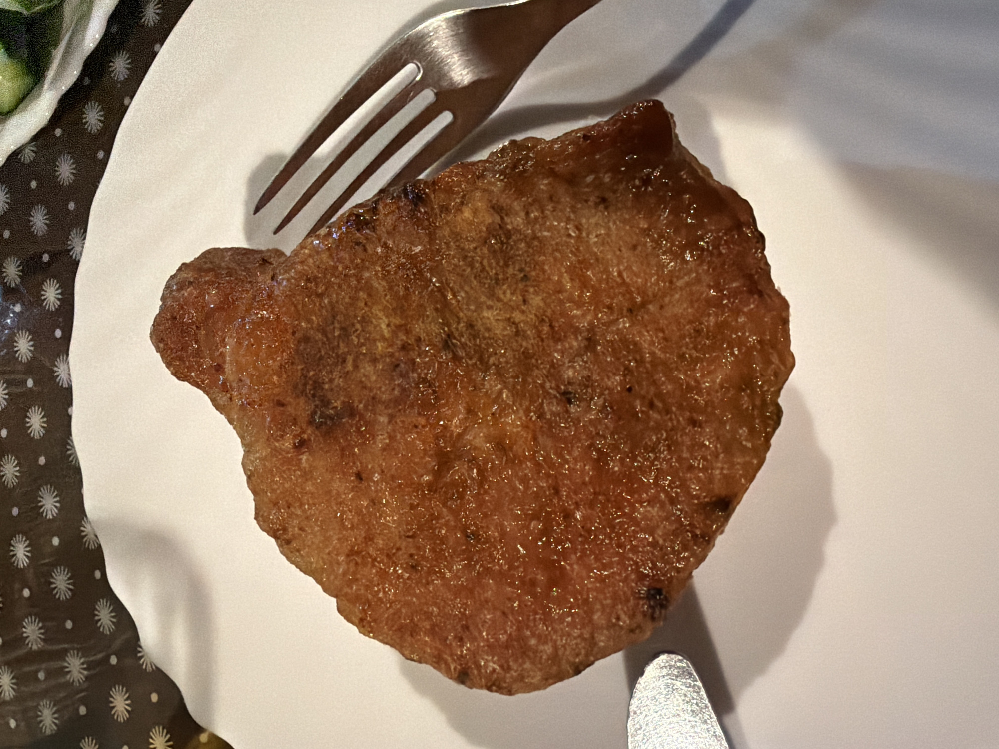 A close-up, top-down view shows a fried piece of meat, possibly a pork chop, centered on a white plate. The meat is a deep golden brown and appears slightly greasy. To the right of the meat, a silver fork with three tines is visible, angled towards the plate. To the left of the meat, a small portion of a silver knife or utensil is also present. The plate is set on a dark brown tablecloth with a pattern of stylized white floral or star shapes. In the upper right corner, out of focus, a plate with green food, possibly salad or vegetables, can be seen. The lighting casts shadows on the white plate, with a pronounced shadow from the meat and fork. The image appears to be taken indoors, likely in a dining setting. There is no discernible location in Sochi, Russia, indicated in the image.
