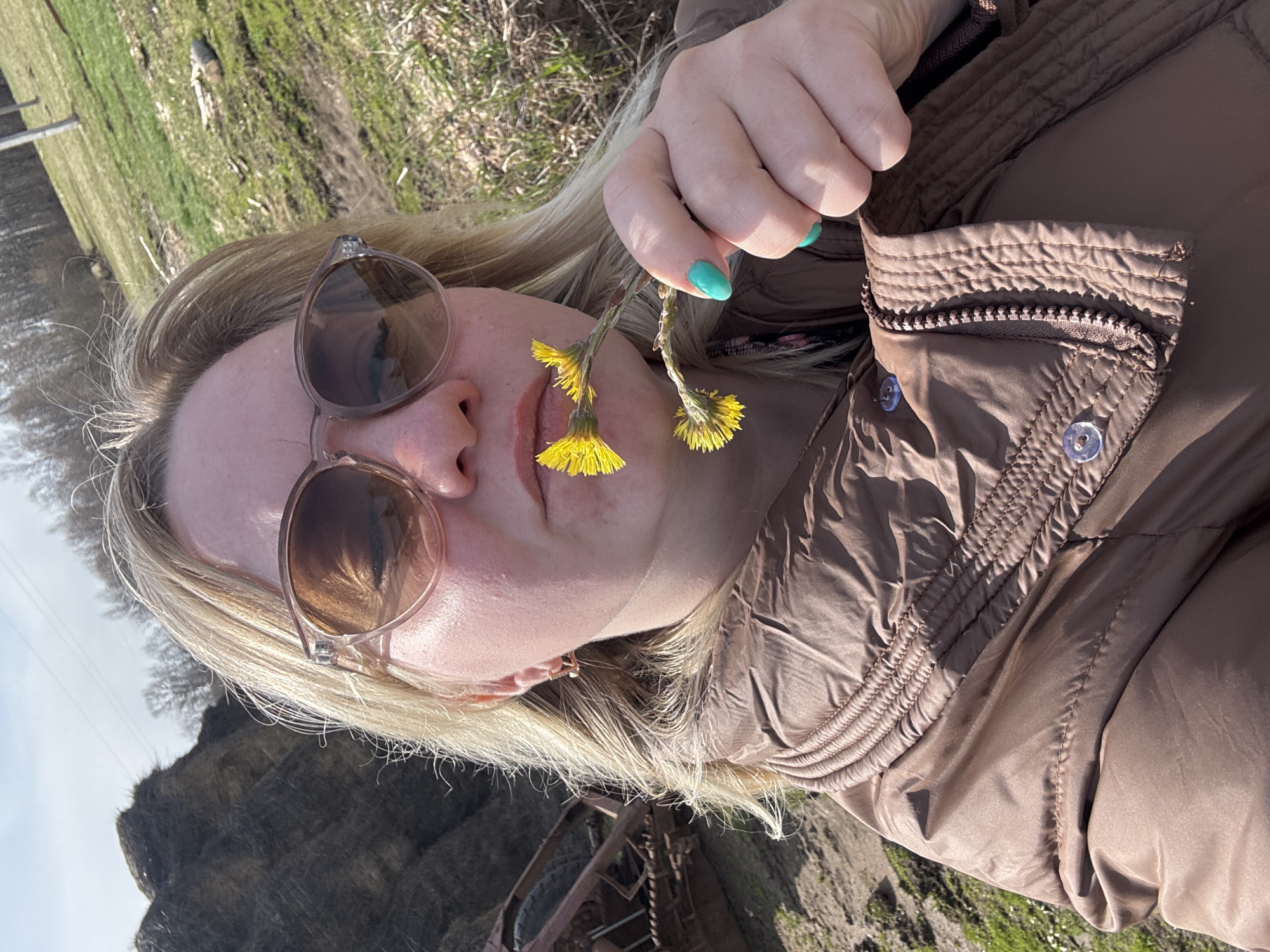 The image shows a person with light blonde hair lying on the ground outdoors, likely on grass. They are wearing sunglasses and a brown quilted jacket, holding a small bunch of yellow flowers near their face. The person has light-colored skin and a visible manicure with teal nail polish. The background consists of a slightly out-of-focus landscape with greenery and what appears to be a weathered, metallic structure.
Based on the location information provided, the scene is likely in a park or open space within the city of Уральское, Russia, though specific landmark identification is impossible from the image alone. The overall impression is that the person is resting in a natural setting.