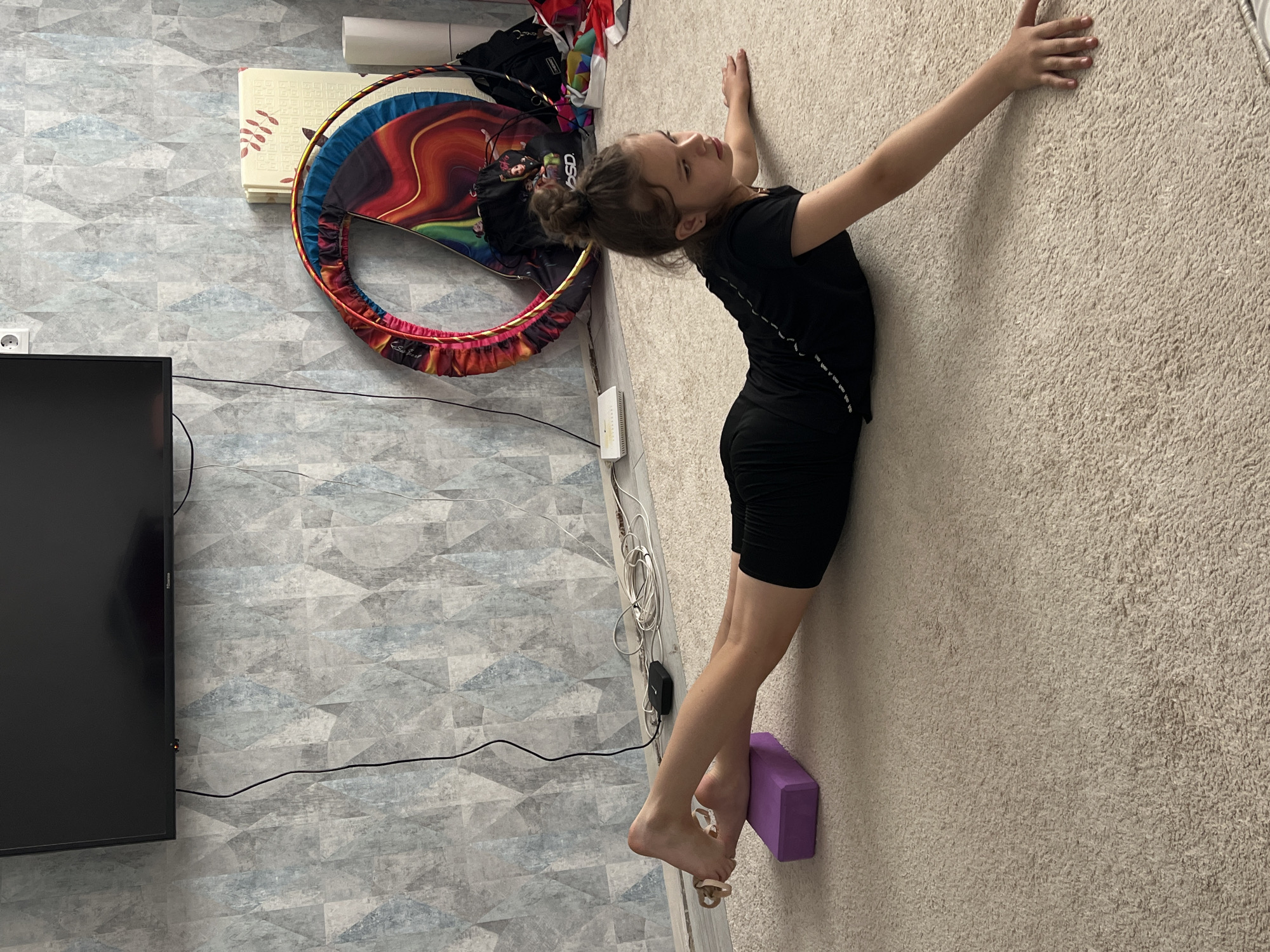 A young girl, likely a pre-teen, with light brown hair styled in a bun, is depicted performing a stretching or flexibility exercise indoors. She wears a black short-sleeved top and black shorts. Her body is slightly arched as she leans against a light-colored, textured wall (possibly carpeted), with her arms extended upwards and pressed against the surface. Her right foot rests on a purple yoga block, assisting in the stretch, and her expression indicates focus and concentration.
The scene is set in an indoor room, characteristic of a residential space in Chaikovsky, Russia. The room features contrasting wall designs; the left side of the frame displays a grey geometric patterned wallpaper, while the area the girl leans against and the floor beneath her are covered in a light beige, highly textured material resembling carpet.
Various household objects are visible: a black wall-mounted television is partially seen on the left. A colorful decorative hoop, possibly for rhythmic gymnastics, with a vibrant fabric cover featuring swirling patterns, leans against the wall behind the girl. Next to the hoop is a black bag, on which the letters "BSD" are clearly visible in white. A white internet router/modem and a white power outlet are situated near the wall, with multiple black and white cables running along the floor and wall. Above the hoop, a light-colored, rectangular object (possibly a folded mat) leans against the wall. The overall lighting suggests it is daytime. Small, faint text, possibly "So Soft," can be seen on the hoop's fabric cover, and a series of small white symbols or text runs down the side seam of the girl's top.