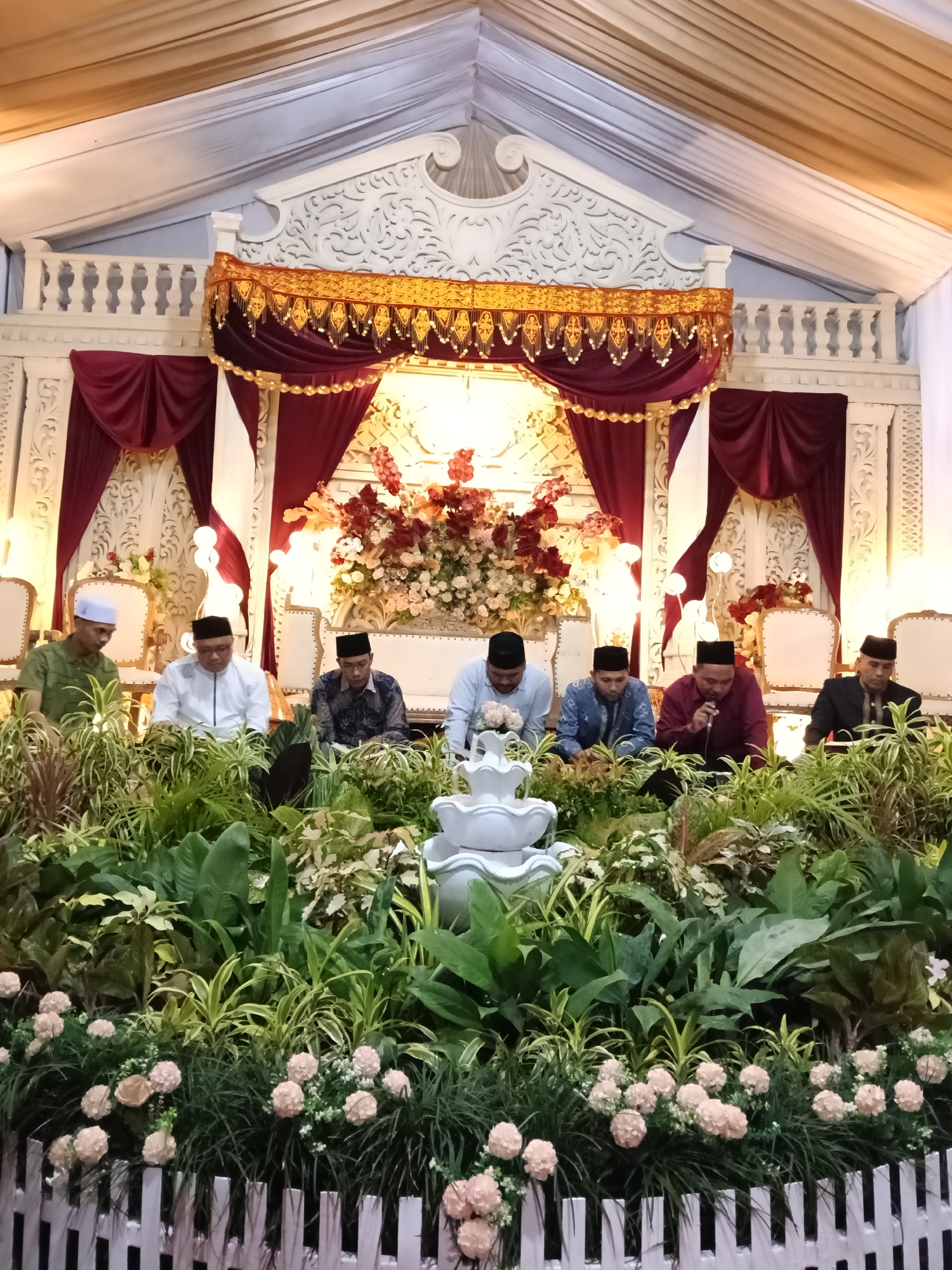 The image shows a ceremonial event in Lhokseumawe, Indonesia, likely a wedding or religious gathering. Seven men are seated in a row, dressed in a variety of formal attire, including traditional caps. They are positioned behind a lush arrangement of greenery and flowers, featuring a tiered fountain. The backdrop is an ornate white structure adorned with red and gold draping. The setting appears to be indoors, possibly in a decorated event space.