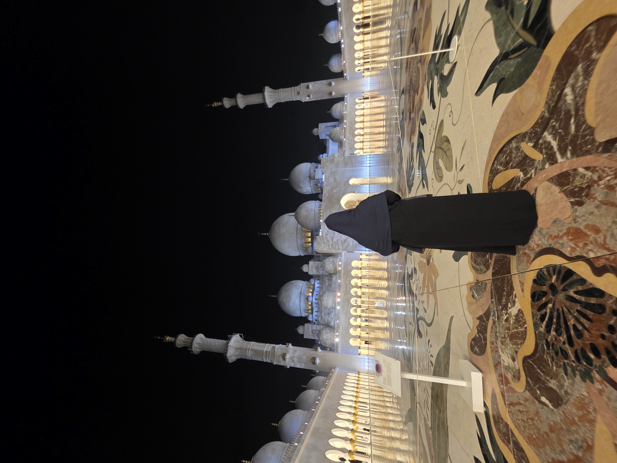 The image shows the Sheikh Zayed Grand Mosque in Abu Dhabi, United Arab Emirates, at night. A woman, viewed from behind, stands on the intricately patterned marble floor of the mosque's courtyard. She is wearing a black abaya and hijab. The mosque's white marble architecture, illuminated by warm lights, is visible in the background, with domes and minarets silhouetted against the dark night sky. The wet floor reflects the lights and the architecture, creating a serene and majestic atmosphere.