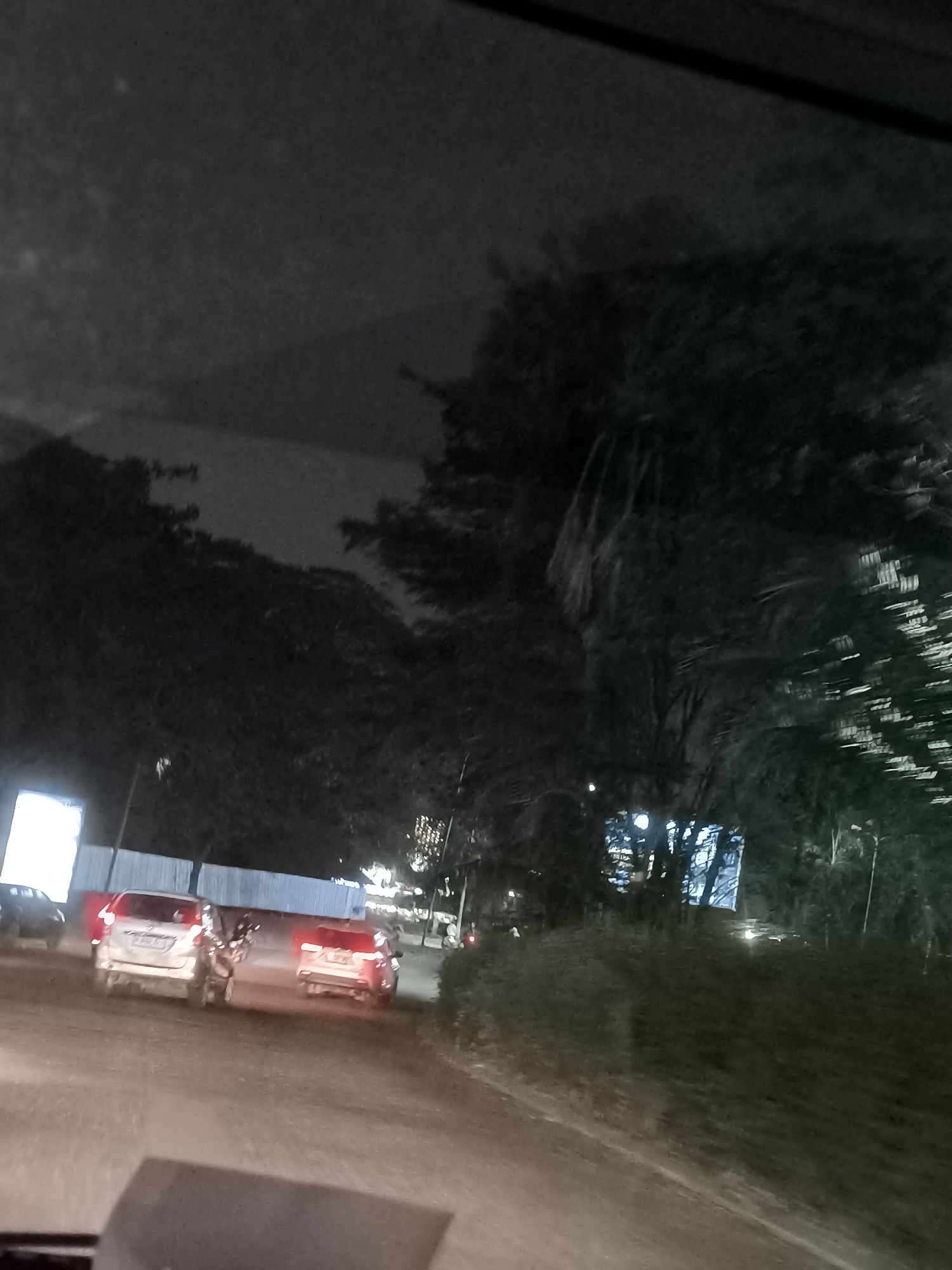 The image depicts a nighttime scene from inside a vehicle in Tangerang, Indonesia, likely on a road under construction or a developing area. In the foreground, the dashboard or car interior is visible. On the road, several cars are present. A white vehicle is ahead, followed by another white vehicle with its brake lights illuminated, indicating it is stopped or slowing down. To the left, another dark-colored vehicle is visible. In the background, large trees create a dark silhouette against the night sky. A large, brightly lit billboard or structure with blue and white elements is situated to the left, behind a temporary fence. There is a sense of ongoing construction or development in the area, suggested by the fence and the unpaved or rough road surface. The lighting conditions are low, with artificial lights from the vehicles and the billboard being the primary sources of illumination.