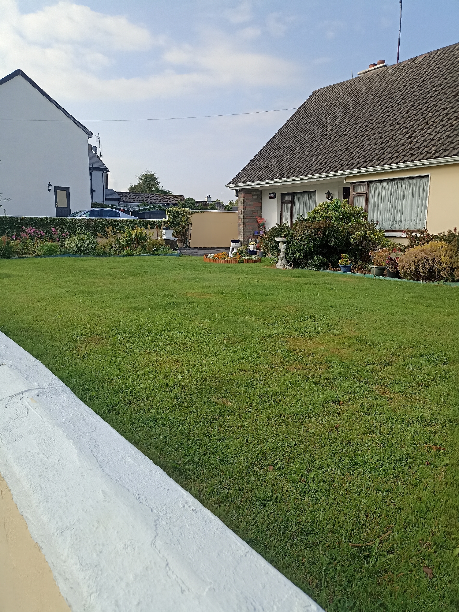The image depicts a residential scene on a clear day in Oughterard, Ireland. In the foreground, a section of a white-painted wall or low barrier is visible, angled diagonally from the lower left to the middle left of the frame. Beyond this, a well-maintained green lawn occupies the majority of the scene.
In the middle ground, a row of well-kept bushes and flowers forms a border along a low wall. Behind this border, a cream-colored single-story house with a dark-tiled roof is visible on the right. The house features several windows with white curtains. Adjacent to this house, to its left, is a white two-story house with a dark-tiled roof and a single dark-colored door. A silver car is parked in front of the white house, behind the floral border. Further in the background, other houses and trees are visible, suggesting a suburban or village setting. A light pole or antenna is present on the roof of the cream-colored house. The sky above is partly cloudy, with patches of blue. The overall impression is one of a quiet, tidy residential area.