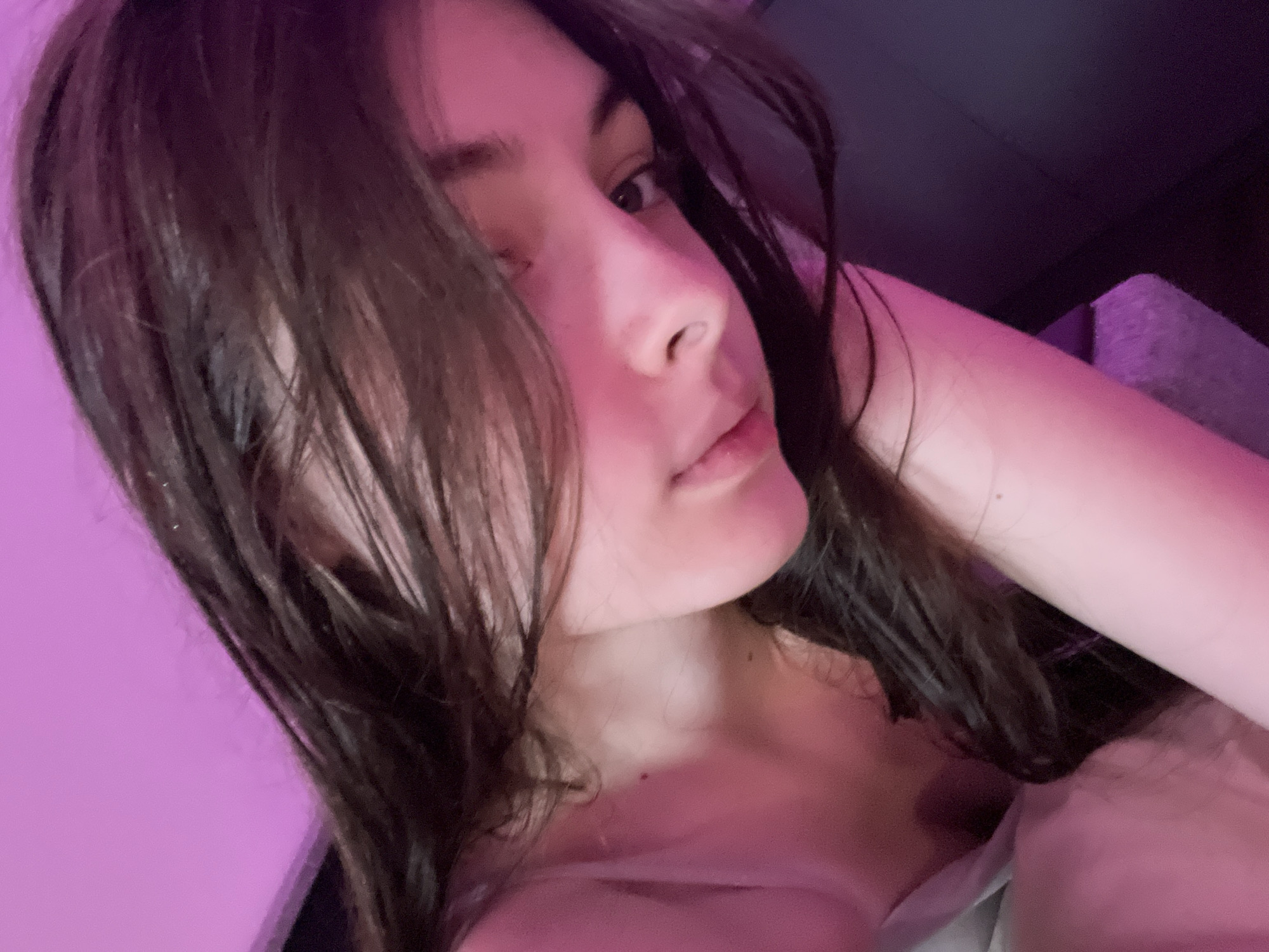 A young woman with long, dark hair is angled towards the camera. The lighting is predominantly pink and purple. The subject's face is partially obscured by her hair. A wall is visible on the left side of the frame. The subject's hand and arm are visible, bent up behind her head. There is some furniture on the right side of the frame. The location in the city of Saint Petersburg, Russia, cannot be determined from this image.