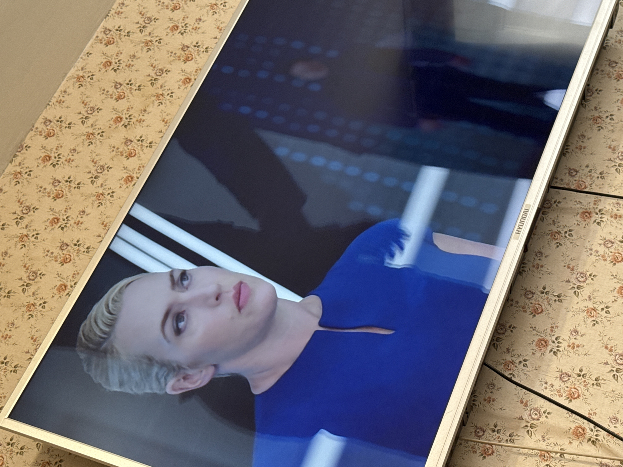 The image shows a Hyundai television displaying a woman with blonde hair wearing a blue dress. The television is placed on a surface covered in floral patterned wallpaper. The location is unknown; the city of Mytishchi, Russia, cannot be confirmed from the image alone.
