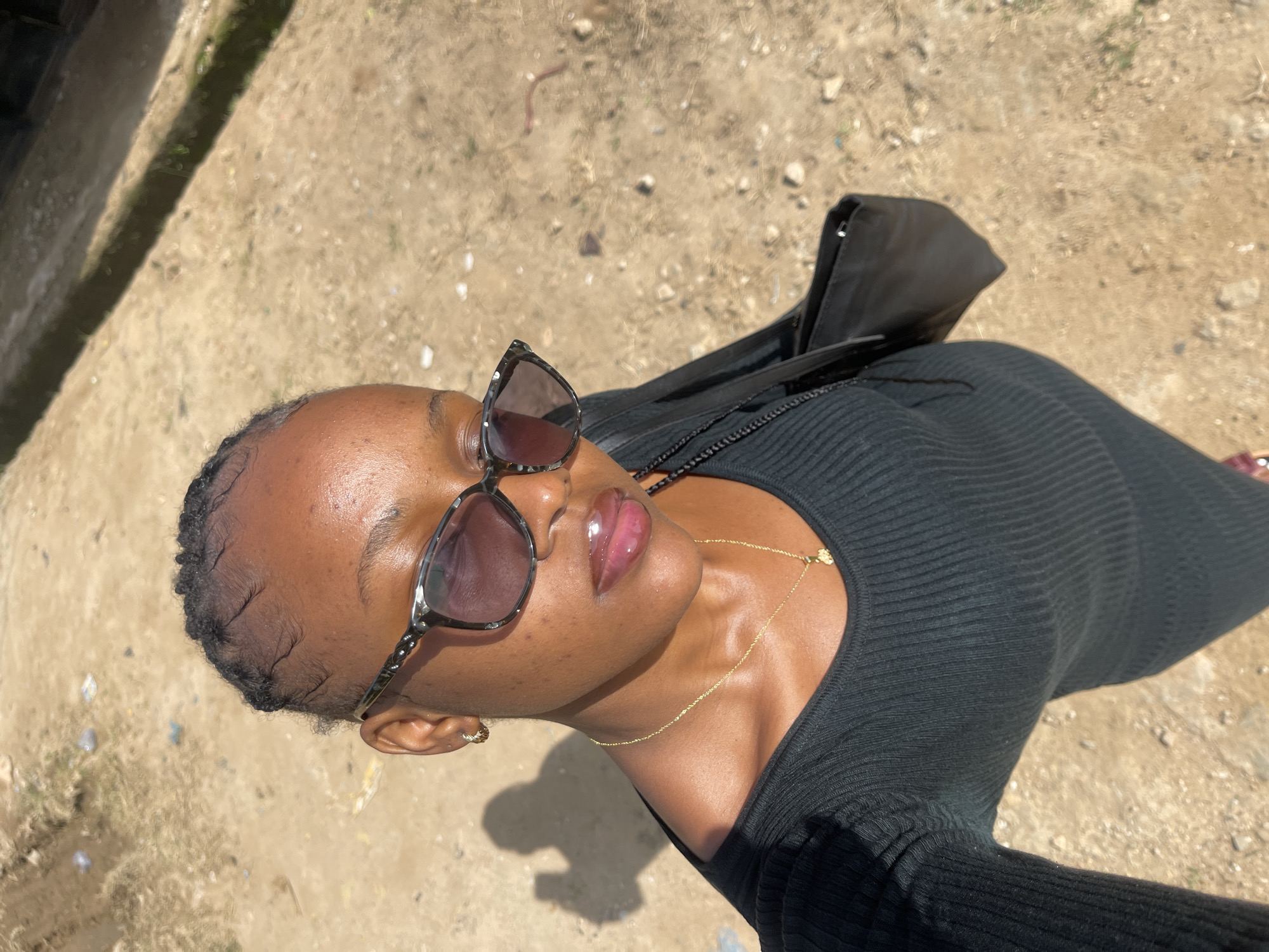 A young Black woman is taking a selfie outdoors, with the sun casting a shadow on the ground to her left. She is wearing a black, long-sleeved, ribbed dress, a gold chain with a pendant, and large, patterned sunglasses. Her hair is braided and pulled back from her face. She has on lip gloss. A black purse is visible on her right side. The ground beneath her appears to be dirt and gravel, with some sparse vegetation and scattered debris. The background is out of focus, showing more of the dirt ground and a hint of a building or structure to the right. This scene could be located anywhere in Jos, Nigeria, as the environment is generic and common to the region.