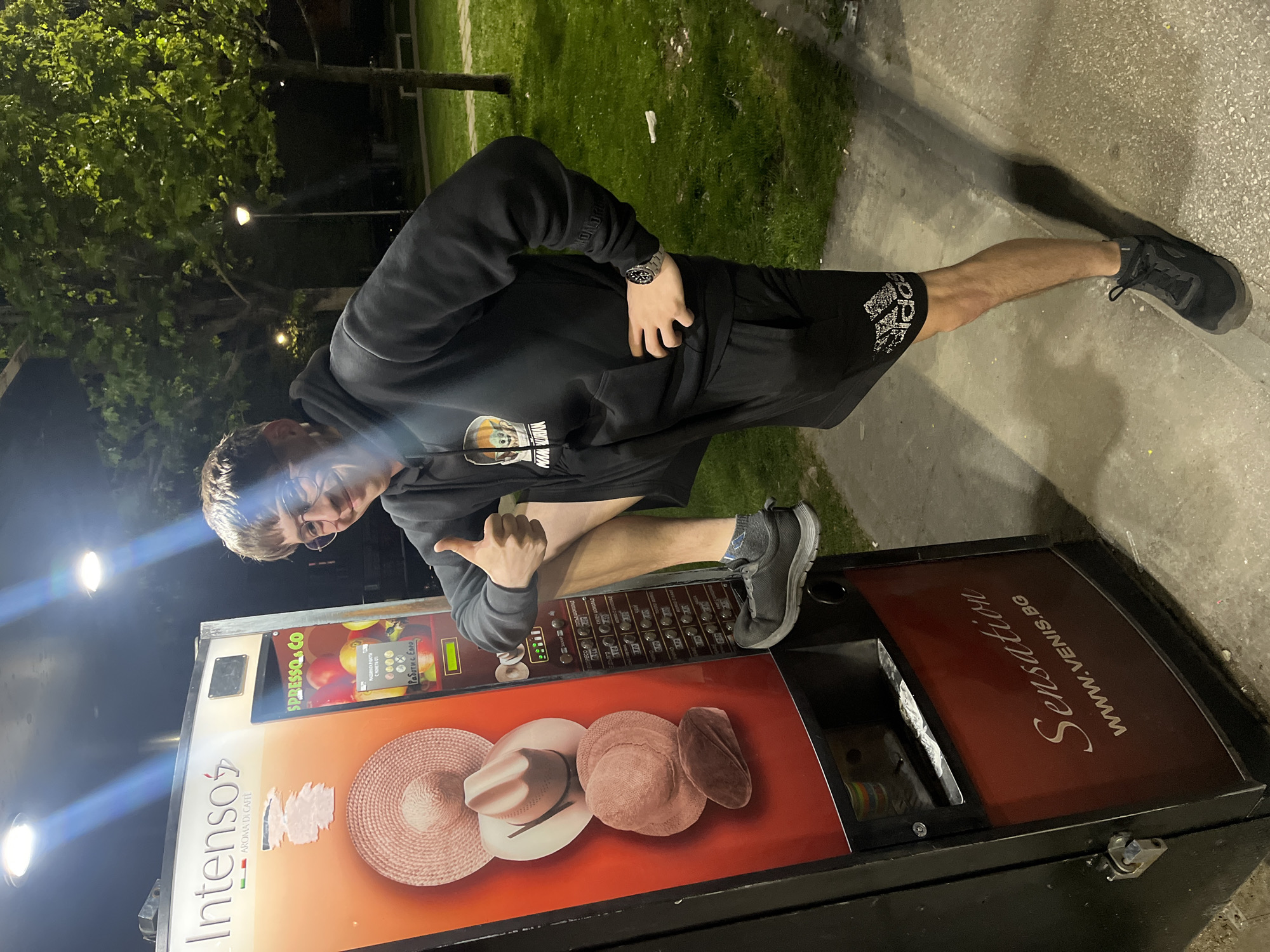 A young man in a dark hoodie, shorts, and sneakers poses beside a vending machine outdoors. He is crouched with his right leg extended onto the machine and is giving a thumbs-up with his right hand. The vending machine, labeled "Intensos Aroma Di Caffe," features images of straw hats and a product selection panel. The location is urban, with green foliage and streetlights visible in the background. The vending machine also advertises "Sensation" and a website, "WWW.VENIS.BG," and displays instructions and prices in a language using Cyrillic script, indicating the location may be in a Slavic country.