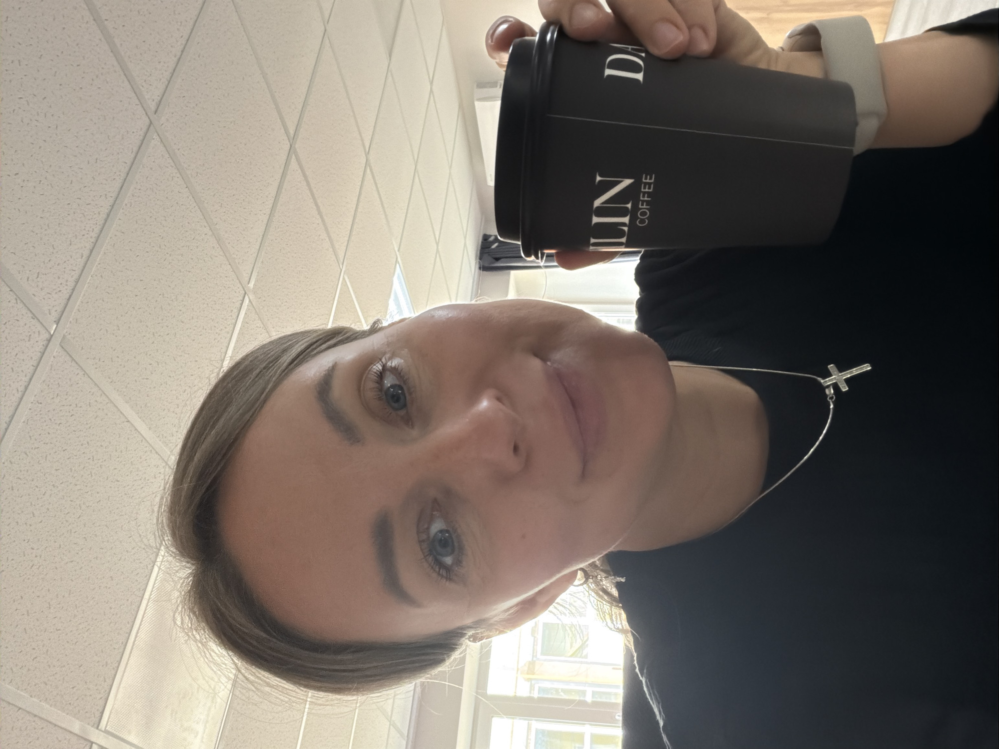 The image is a selfie taken by a woman, oriented vertically with the top of her head towards the bottom of the frame. She looks directly at the camera with a neutral to slightly smiling expression, featuring blue eyes and light brown hair. She wears a black top and a silver necklace with a cross pendant.
Her hand holds a black disposable coffee cup, positioned in the upper right quadrant of the image. Visible white text on the cup clearly reads "LIN COFFEE" and partially "DA", suggesting a brand like "DARLIN COFFEE" or "DAHLIN COFFEE".
The background primarily shows a light-colored drop ceiling with square tiles, characteristic of an indoor commercial or office space. A window is visible behind the woman, showing daylight and blurred buildings outside. The overall lighting is bright, indicating daytime. The scene is set in what appears to be an interior location in Chaikovsky, Russia.