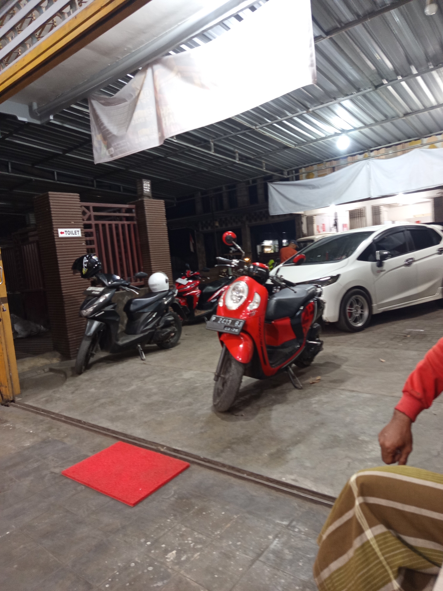 The image depicts an outdoor scene in Prajekan Lor, Indonesia, likely a parking area or a covered walkway adjacent to a building. Several motorcycles are parked, including a black scooter with a white helmet on its handlebars and a red scooter in the foreground. A white car is also visible parked behind the red scooter. A sign with an arrow pointing to "TOILET" is mounted on a brick structure. The environment appears to be dimly lit, suggesting it might be evening or nighttime. A red mat is placed on the ground in front of the motorcycles. The presence of a person's arm in a red sleeve and a patterned garment in the lower right corner suggests human presence, though the individuals are not fully visible. The scene is characterized by a mix of vehicles, infrastructure, and a sense of everyday activity in a localized area.