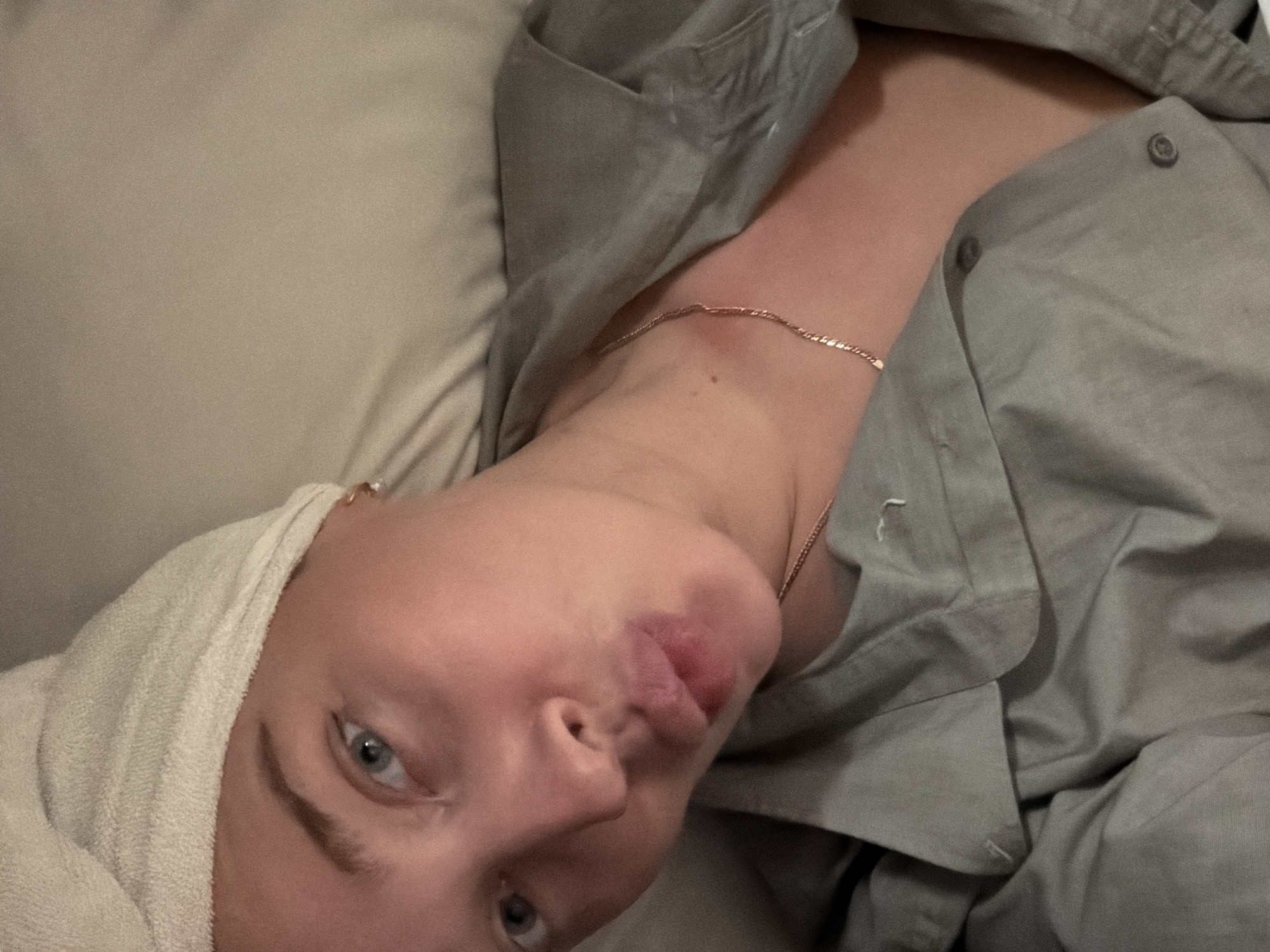 The image depicts a woman lying down, likely in a bed or on a couch. She has a white towel wrapped around her head and is wearing a light gray button-down shirt. A gold necklace is visible around her neck. The background appears to be a light-colored pillow or cushion. The location in Moscow, Russia, is unknown.