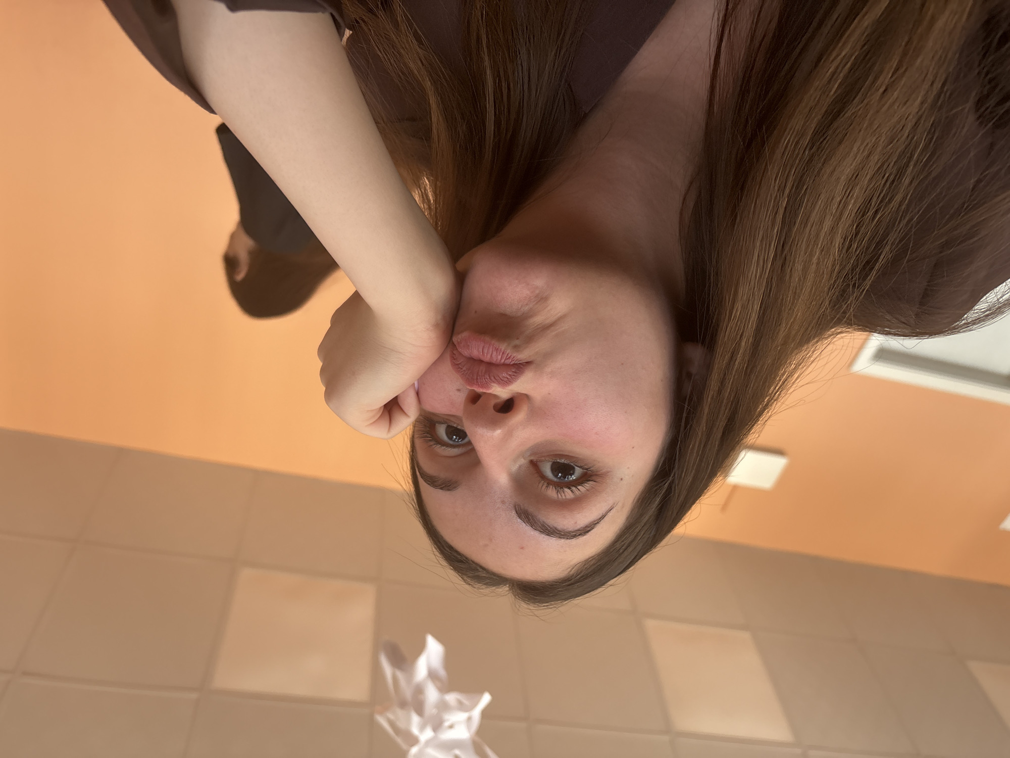 A woman with fair skin and brown hair is positioned in the foreground of the image. She is resting her cheek on her hand and pursing her lips slightly, seemingly posing for a selfie. The background shows a tiled wall and ceiling in a peach/beige hue, possibly indicating a bathroom or similar interior space. Given the prompt, the location is potentially Burgas, Bulgaria, but there are no specific indicators of location visible in the image. There is also a crumpled, white object, possibly a tissue, in the lower part of the frame.