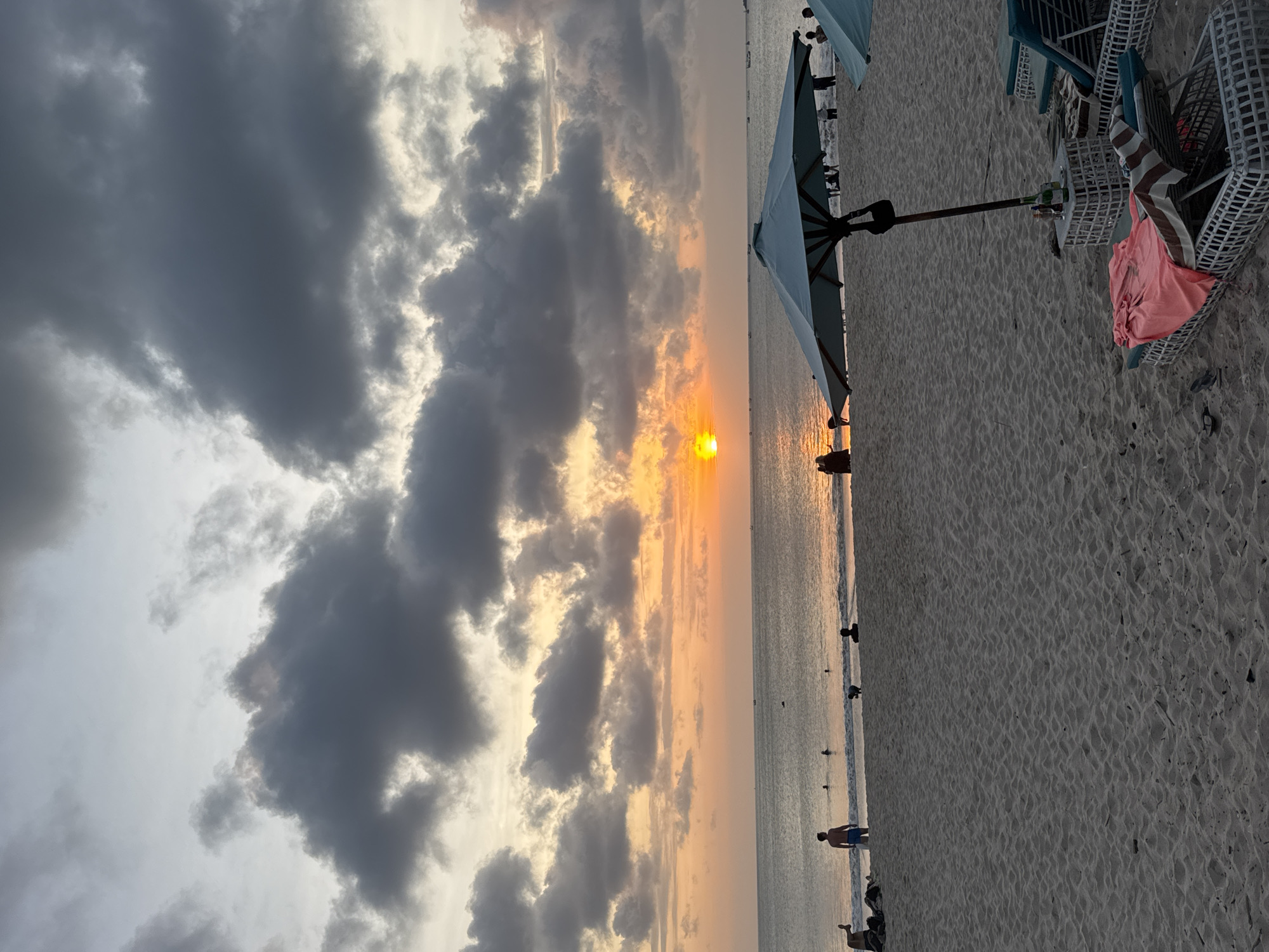 This image depicts a sunset scene at a beach in Kuta, Indonesia. The photograph, taken in a vertical orientation, shows a sandy beach with several beach chairs and umbrellas set up. The ocean stretches out to the horizon, where a vibrant sunset is occurring. The sky is filled with dramatic, dark clouds illuminated by the orange glow of the setting sun.
Several people are visible on the beach and in the shallow water, appearing to enjoy the evening. Some are standing and walking along the shoreline, while others are partially submerged in the water. A few beach chairs are arranged on the sand, some with towels draped over them, suggesting a relaxing atmosphere. The sand itself is finely textured and appears to be typical of a tropical beach.
The time of day is clearly sunset, indicated by the warm colors in the sky and the low angle of the sun. The weather appears to be a mix of cloudy and clear, with the clouds adding a dramatic element to the sunset. The location cues, such as the beach chairs, umbrellas, and the overall setting, are consistent with a popular beach resort area like Kuta. The overall mood conveyed by the image is one of tranquility and natural beauty. There is no visible text in the image.
