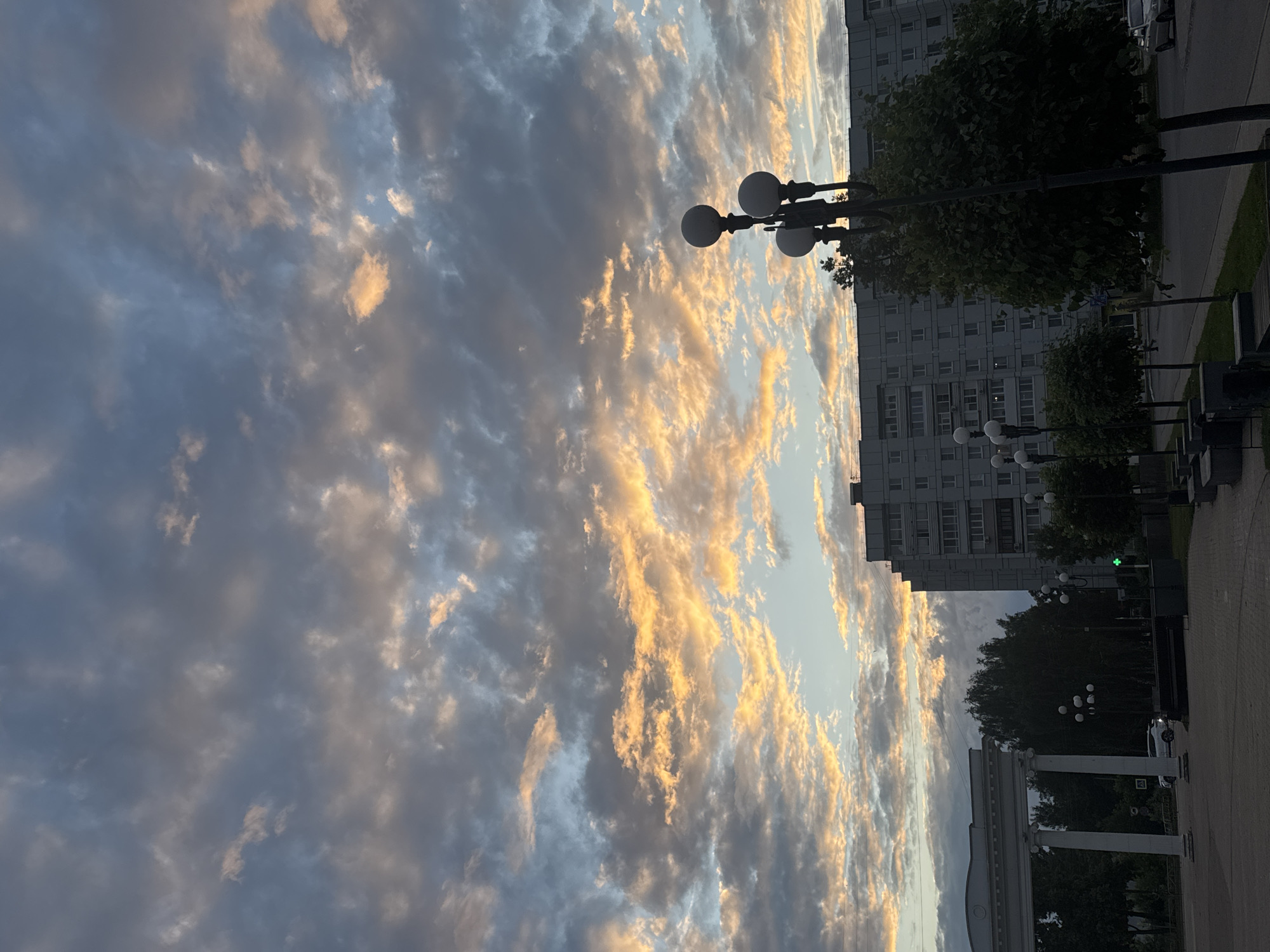 The image depicts a sky filled with dramatic clouds tinged with golden and orange hues, suggesting either sunrise or sunset. Below the sky, a typical Soviet-era apartment building dominates the mid-ground. To the left, a white classical-style archway with columns stands out, possibly marking an entrance or a public space. In the foreground, a street with a sidewalk is visible, lined with trees and streetlights. One lamppost, with multiple globe-shaped lights, is prominently positioned in the right foreground. Cars are present on the street, and a pedestrian crossing light with a green signal is visible. The overall scene appears to be a public square or street in a city, likely Chaikovsky, Russia, given the architectural style of the buildings.
