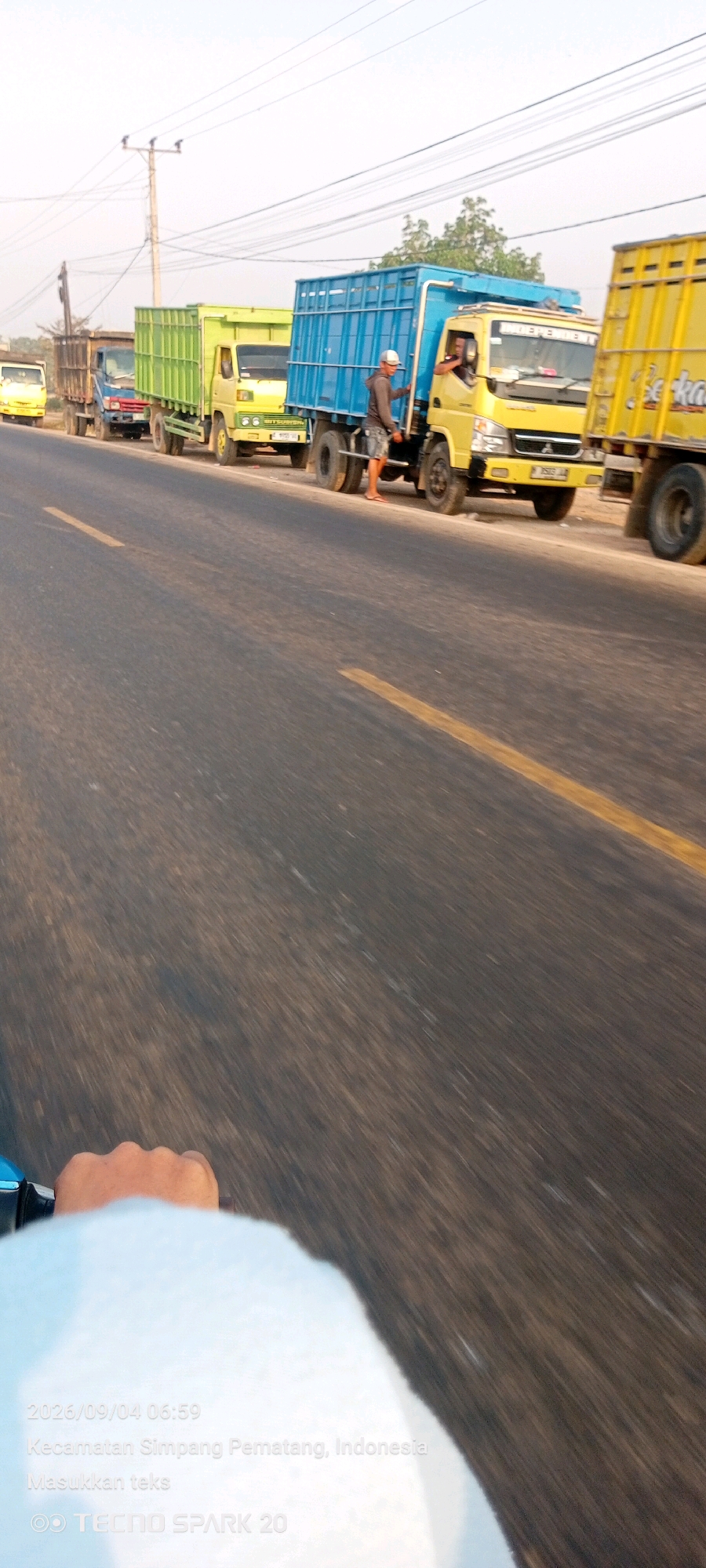 The image shows a line of trucks parked on the side of a road in Simpang Pematang, Indonesia. The trucks are of various colors, including yellow, green, and blue. Some of the trucks appear to be loaded. There are a few people visible, including a person wearing a white hat and dark clothing standing next to a blue truck, and another person in the driver's seat of a yellow truck. The road is paved with a yellow line marking the lane. The sky is overcast. The image is dated 2026/09/04 06:59. The lower portion of the image shows a blurry view of a hand and part of a person's head, suggesting the photo was taken from a moving vehicle, likely a motorcycle. The text "Kecamatan Simpang Pematang, Indonesia" and "Masukkan teks" are overlaid on the image, along with "TECNO SPARK 20".