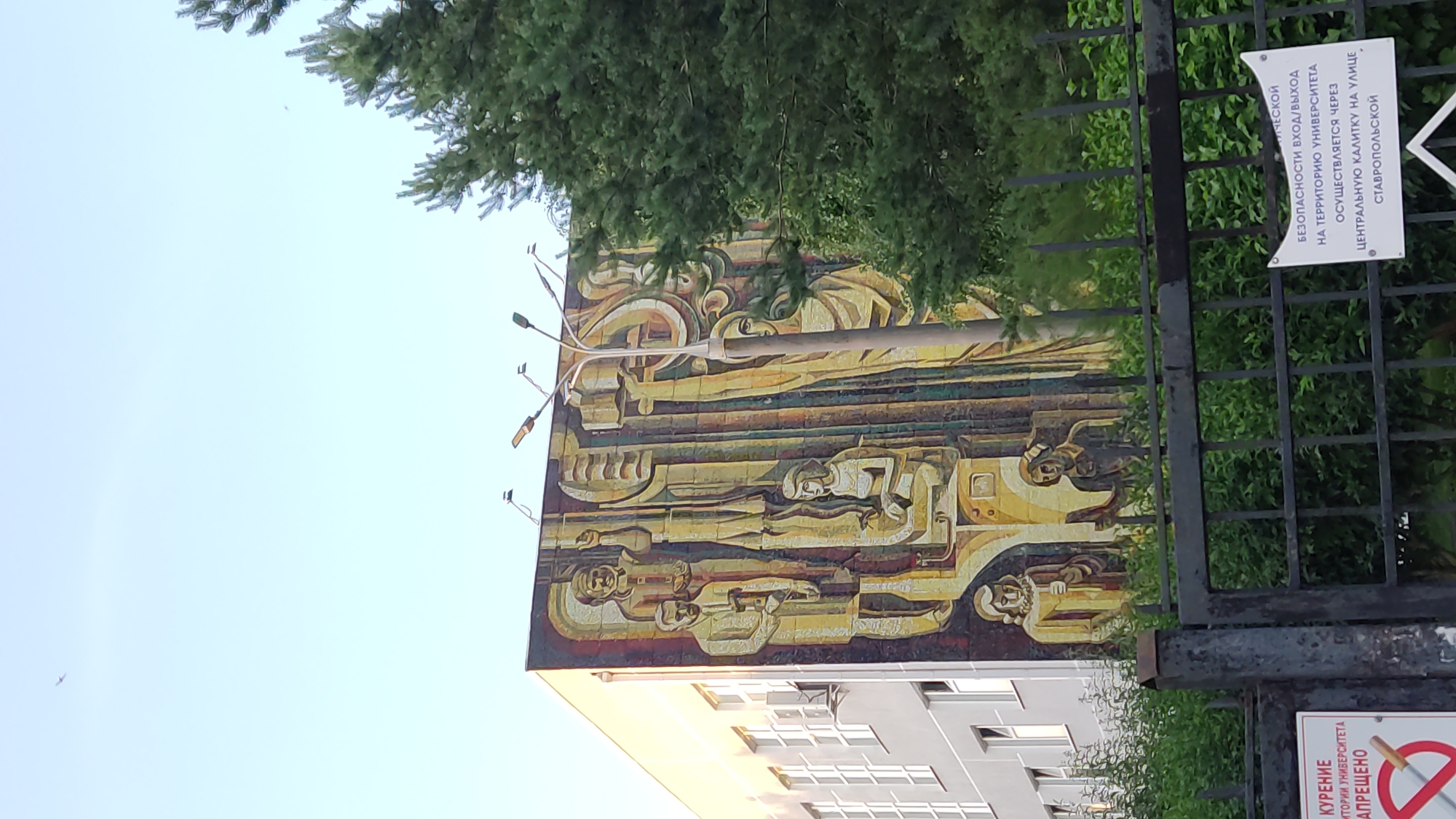 This media file is a photograph taken from a low angle, looking upwards. The primary subjects are a large, colorful mural on the side of a building and a black metal fence in the foreground. The mural features abstract figures and geometric shapes in shades of yellow, brown, red, and black. It appears to depict a narrative or theme, possibly related to progress or community, with stylized human forms integrated into the design. Above the mural, green foliage from a tree partially obscures the sky and the upper portion of the building.
The building itself is modern, with visible windows and balconies suggesting an urban setting, likely an apartment building or institutional structure. Attached to the fence is a white sign with text in Russian, indicating rules or information for the location. The sign reads: "SMOKING IS PROHIBITED ON THE UNIVERSITY GROUNDS. ENTRY/EXIT IS THROUGH THE CENTRAL GATE ON STAVROPOLSKAYA STREET." This text provides a crucial clue about the location, identifying it as part of a university campus on Stavropolskaya Street, and that the photograph was taken in Krasnodar, Russia.
The lighting suggests it is daytime, likely morning or afternoon, with bright but slightly overcast conditions that contribute to the soft illumination of the scene. No specific activity or event is clearly depicted, but the mural itself suggests a historical or artistic commemoration. The overall impression is one of an urban environment with elements of public art and regulatory signage. The perspective emphasizes the scale of the mural and the imposing nature of the fence.