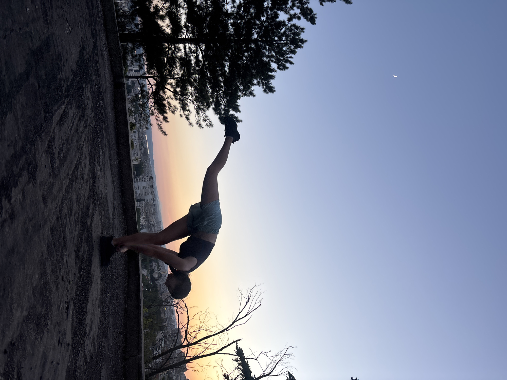 A silhouette of a woman is captured in a yoga pose, stretching forward with one leg extended behind her. She is on a dark, textured surface, possibly a rooftop or overlook, with a city skyline visible in the background. The sky is a gradient of soft blues and oranges, indicating either dawn or dusk. A crescent moon is visible in the upper left portion of the sky. Trees, one a dark evergreen and the other bare branches, frame the scene. The location appears to be an elevated vantage point within the city of Varna, Bulgaria, offering a panoramic view of the urban landscape.