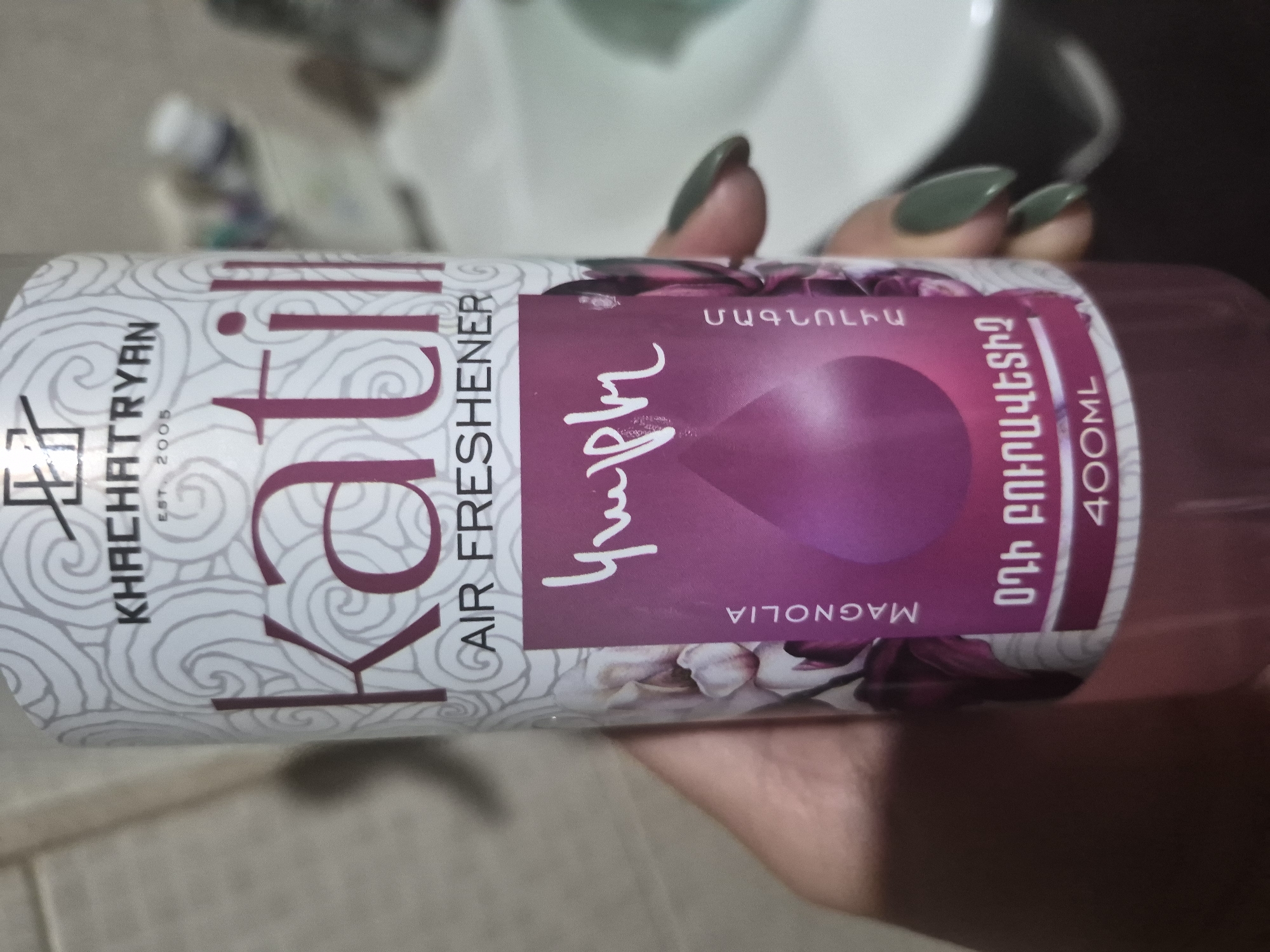 The image displays a close-up of a hand holding an aerosol can, likely an air freshener, in an indoor setting. The hand, featuring long nails painted a dark olive green, holds the can upright.
The can itself is predominantly white with a swirling, decorative pattern. A large purple section adorns the right side, featuring a faint image of magnolia flowers. Visible text includes the brand "KHACHATRYAN" with "EST. 2005" below it. The product name "Kafili" (or "Kafi") is prominent, followed by "AIR FRESHENER". Within the purple section, the scent "MAGNOLIA" is written both vertically in English and horizontally in Armenian script ("ՄԱԳՆՈԼԻԱ"). Below the Armenian for Magnolia, the text "ՕԴԻ ԲՈՒՐՎԱՆԵԼԻՉ" is visible, translating to "Air Freshener". A stylized, handwritten script that appears to be "կաֆի" (Kafi) in Armenian is also present on the purple background. The volume "400ML" is clearly indicated.
The background is out of focus, showing blurred light-colored surfaces, typical of an indoor home environment. The activity is simply holding the product. The presence of Armenian text on the can aligns with the provided context of Yerevan, Armenia, suggesting the product's origin or market.