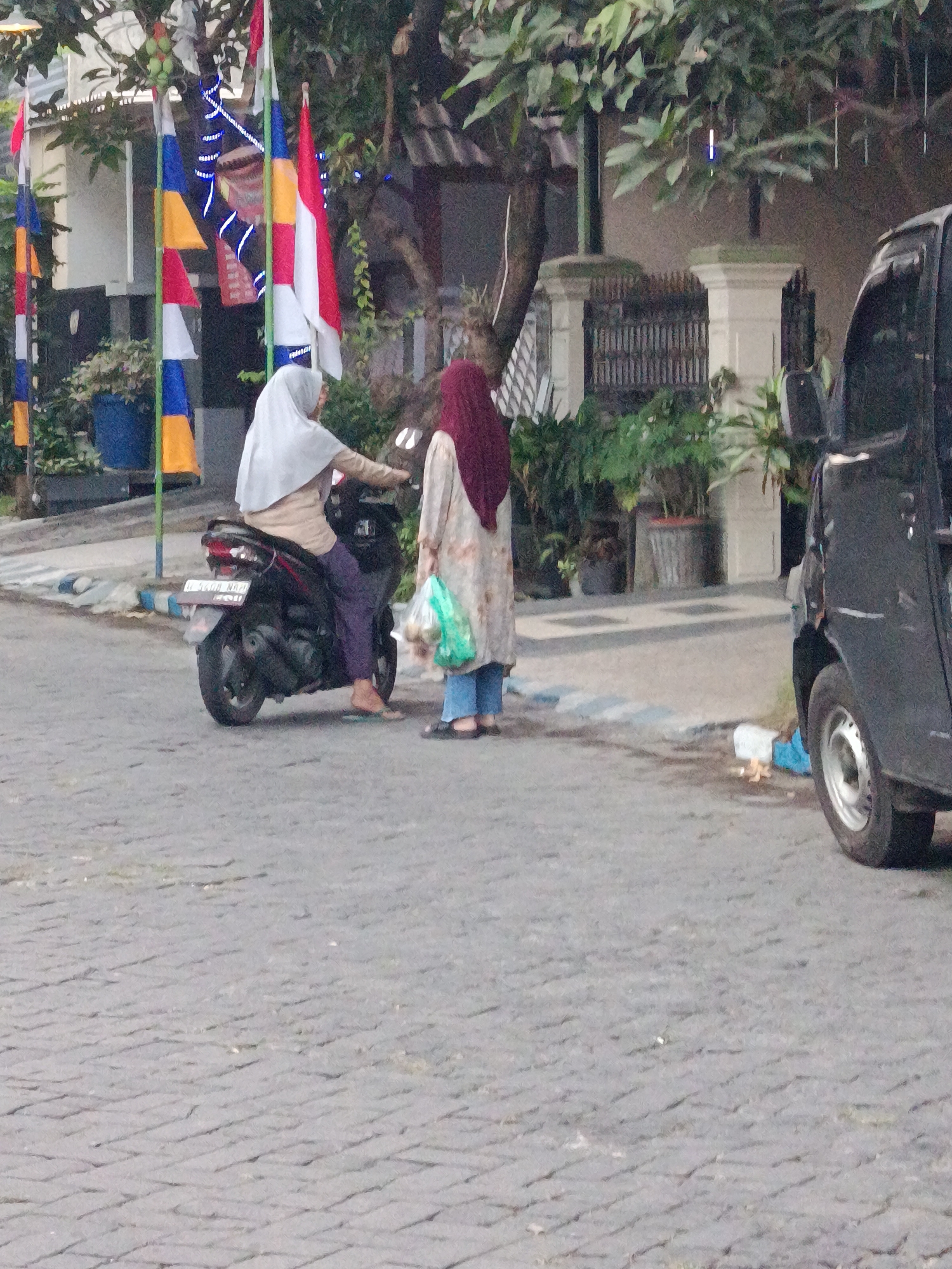 In the city of Keboansikep, Indonesia, a street scene unfolds. A woman, wearing a light gray hijab and a tan top, is seated on a black motorcycle. Her legs are clad in dark purple pants, and her feet are bare, with one sandal visible. Beside her, a second woman stands, holding a light green plastic bag containing what appears to be groceries. She is wearing a maroon hijab and a light-colored patterned garment over blue jeans. A black, three-wheeled vehicle is parked on the right side of the image, partially obscuring the background. The ground is paved with gray interlocking stones. In the background, trees with green foliage are visible, along with a white building with a dark-colored gate. Several colorful flags, red, white, blue, and yellow, are mounted on poles, some adorned with blue lights.